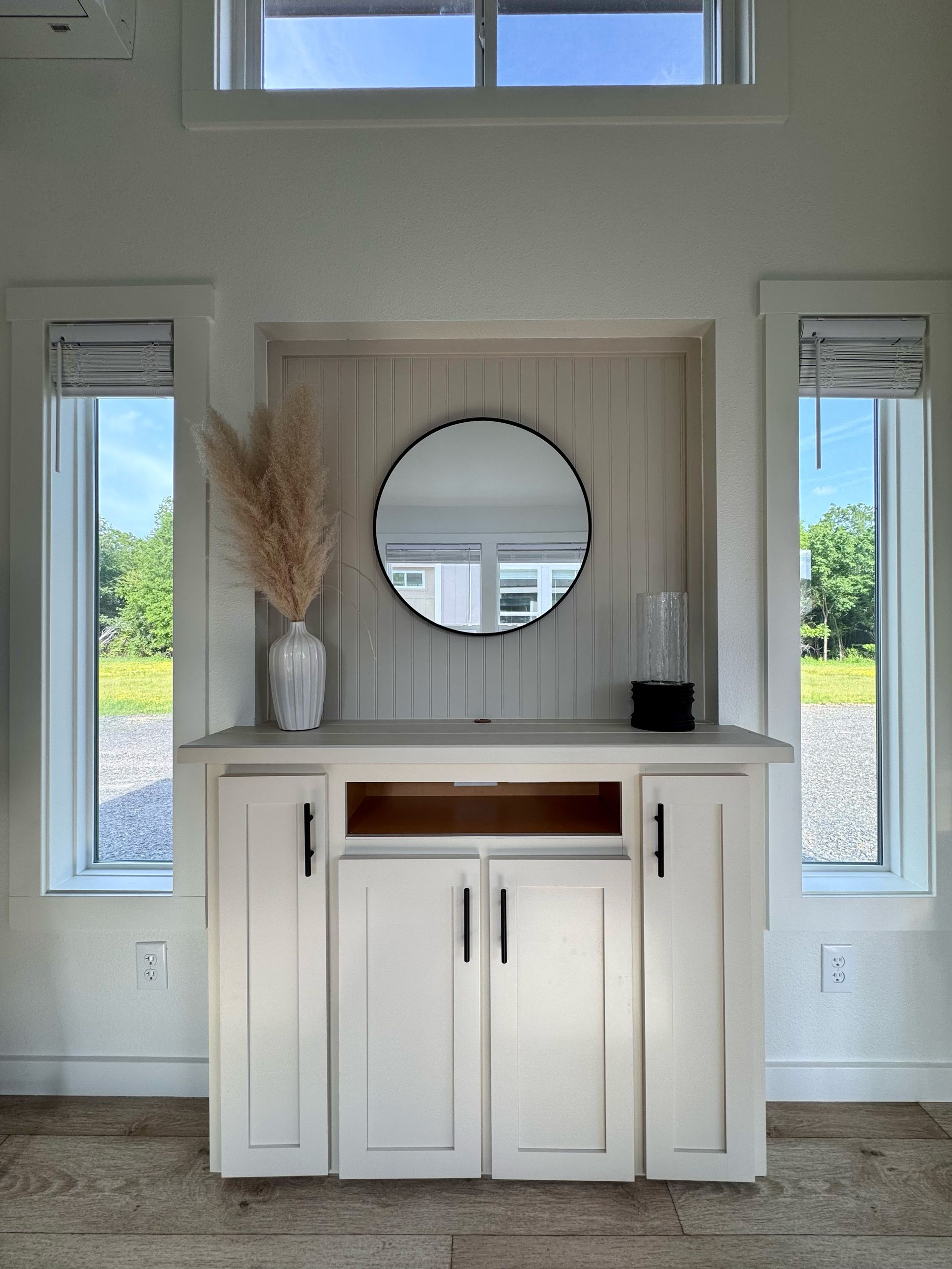 White cabinet with a round mirror, pampas grass, and windows. Interior view.