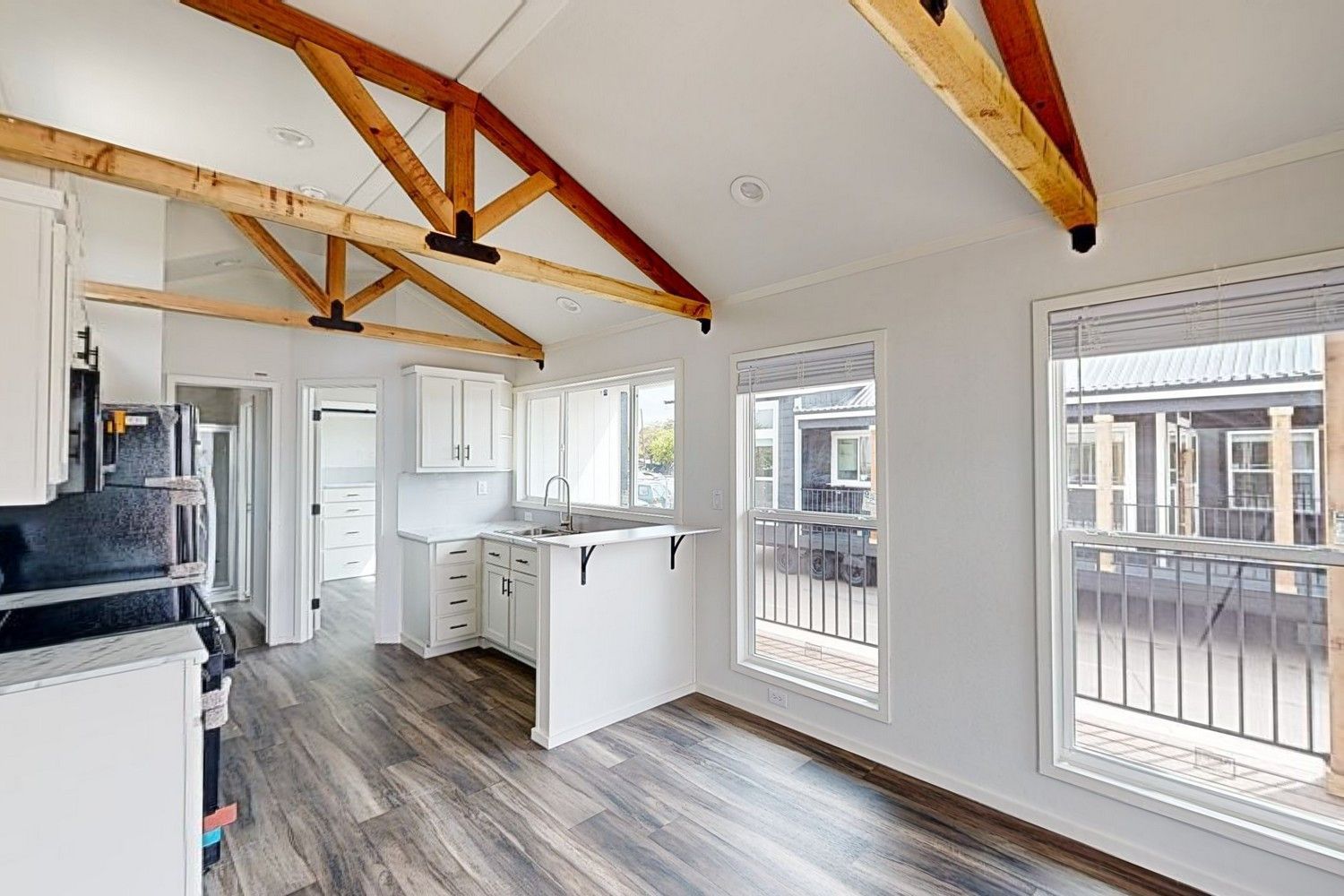 Interior of a small white kitchen and living space, with wooden beams on the ceiling, two windows, and dark flooring.