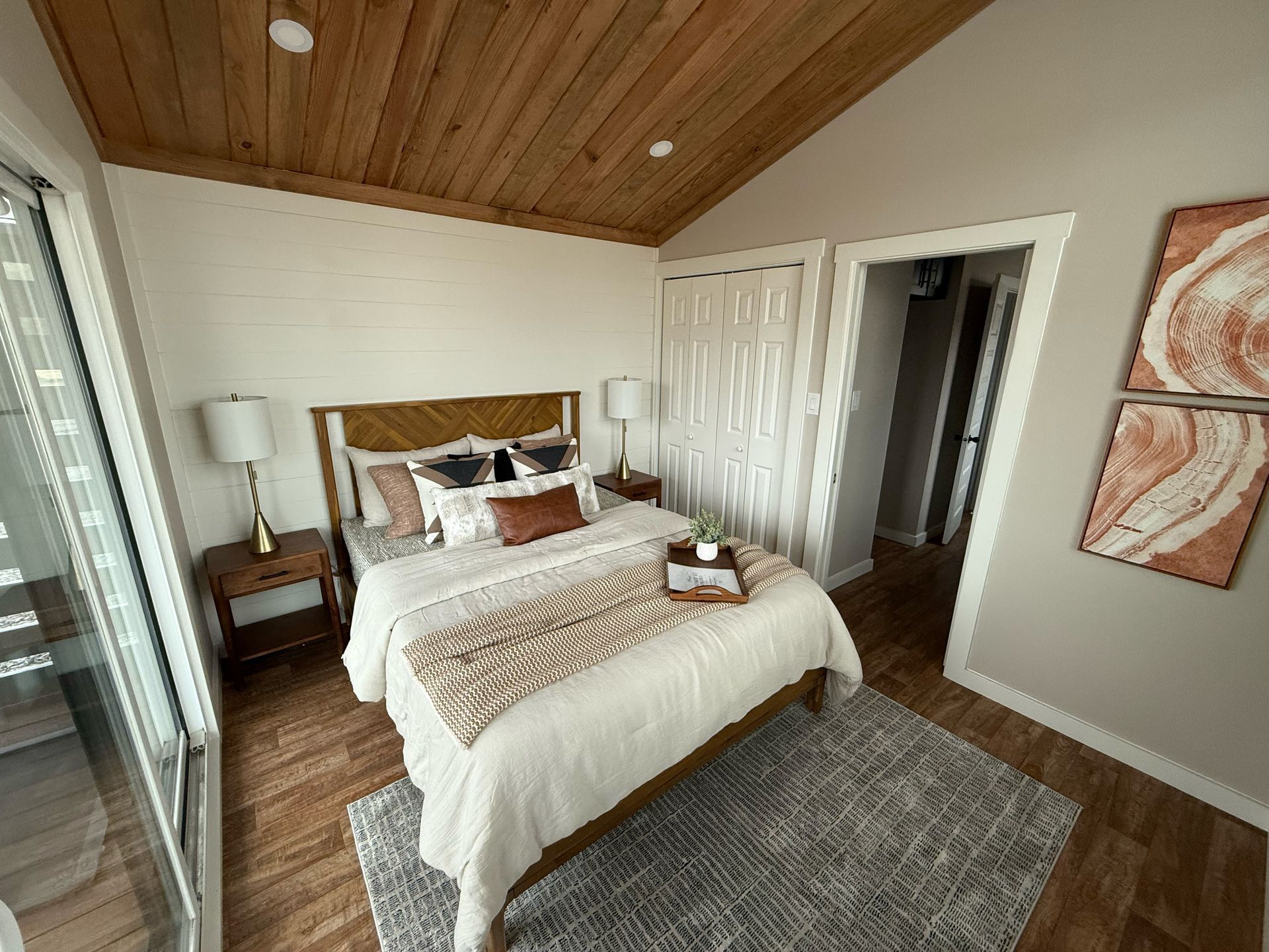 Bedroom with wooden ceiling, neutral walls, bed with pillows, rug, side tables, and artwork.