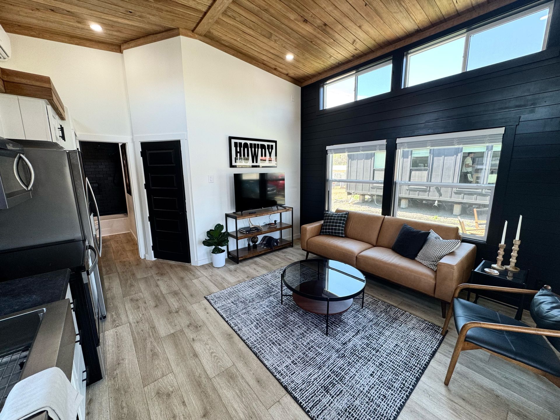 Modern living room with black accent wall, tan sofa, wooden ceiling, rug, coffee table, and windows.