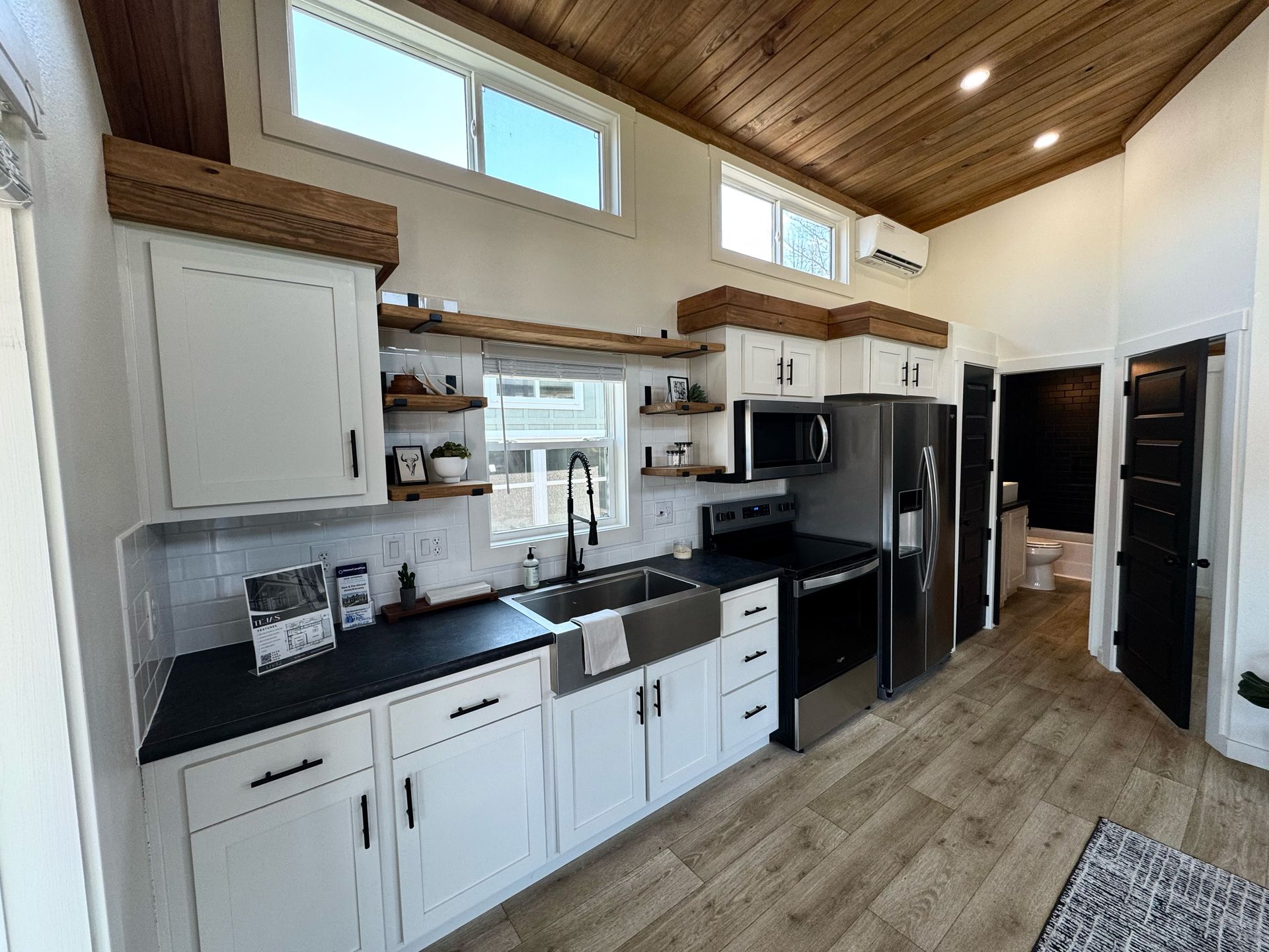 Tiny house kitchen with white cabinets, dark countertops, stainless steel appliances, and wood ceiling.