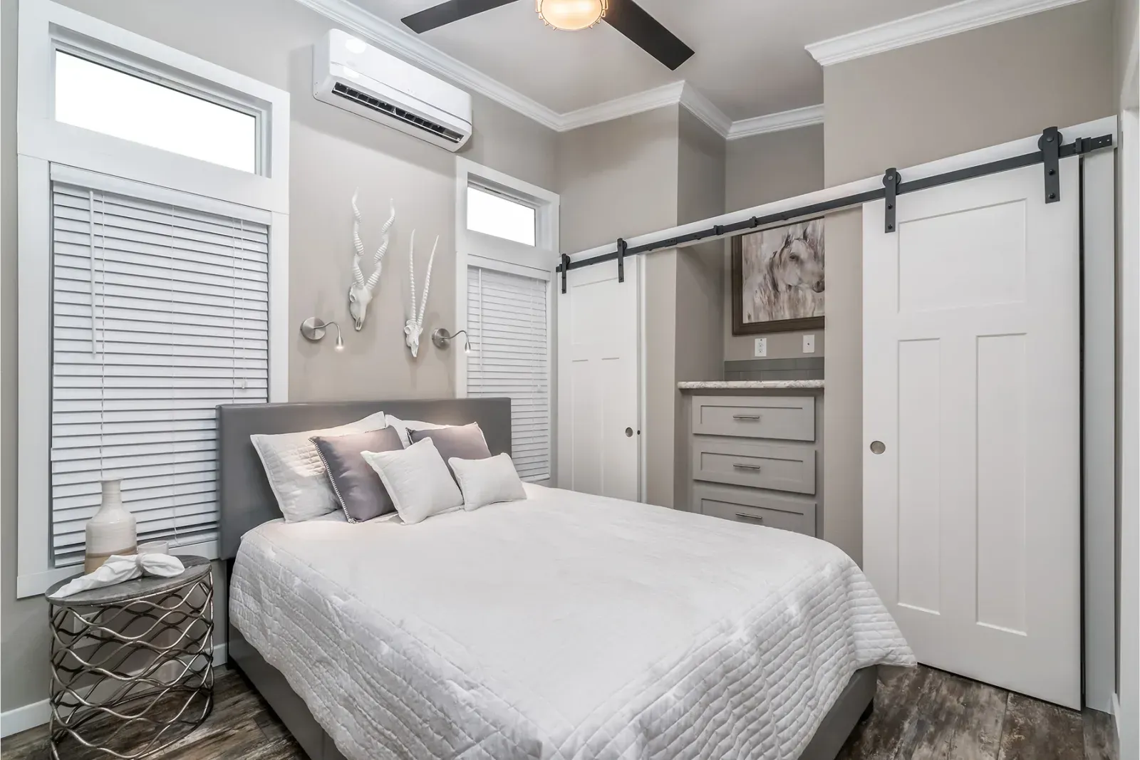 Bedroom with a gray bed, white bedding, and light gray walls. Includes a dresser, closet doors, and windows.