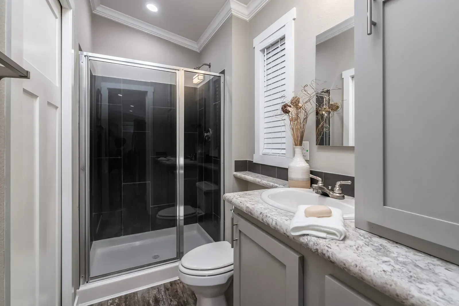 Bathroom with a glass-enclosed shower, gray cabinets, white sink and toilet, and a window with blinds.