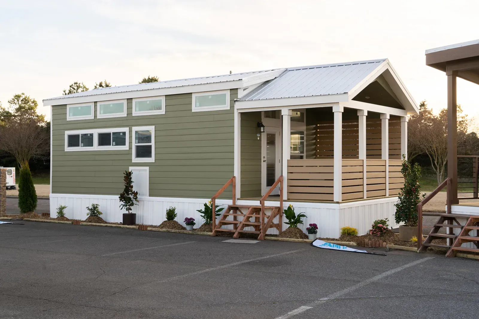 Green tiny house with white trim, porch, and metal roof in a parking lot.