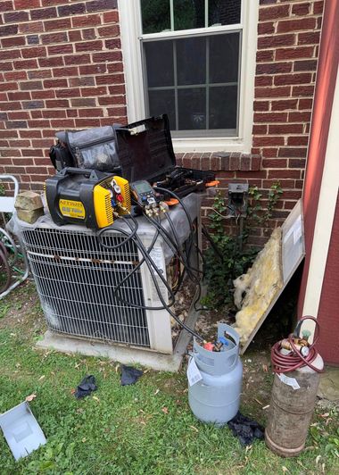 Two outdoor HVAC units beside a brick house, one with tools and debris on top, near a window and grass.