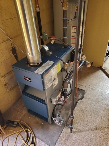 Blue furnace with metal ducts and pipes in a utility room, beside a concrete floor and yellow wall.