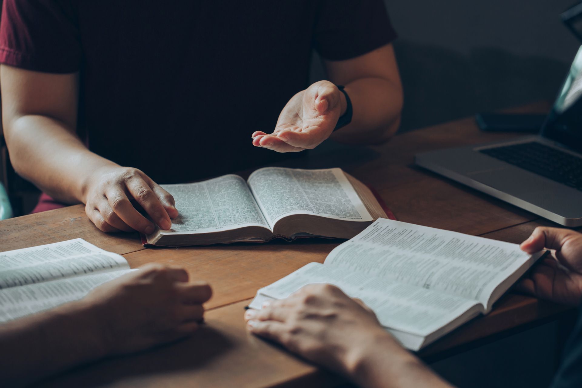 Three people at a table studying open books, one gesturing with hand. A laptop sits nearby.