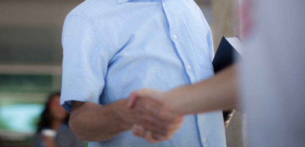 Two people shaking hands, blurred background. One person wears a blue button-up shirt, holding a dark object.