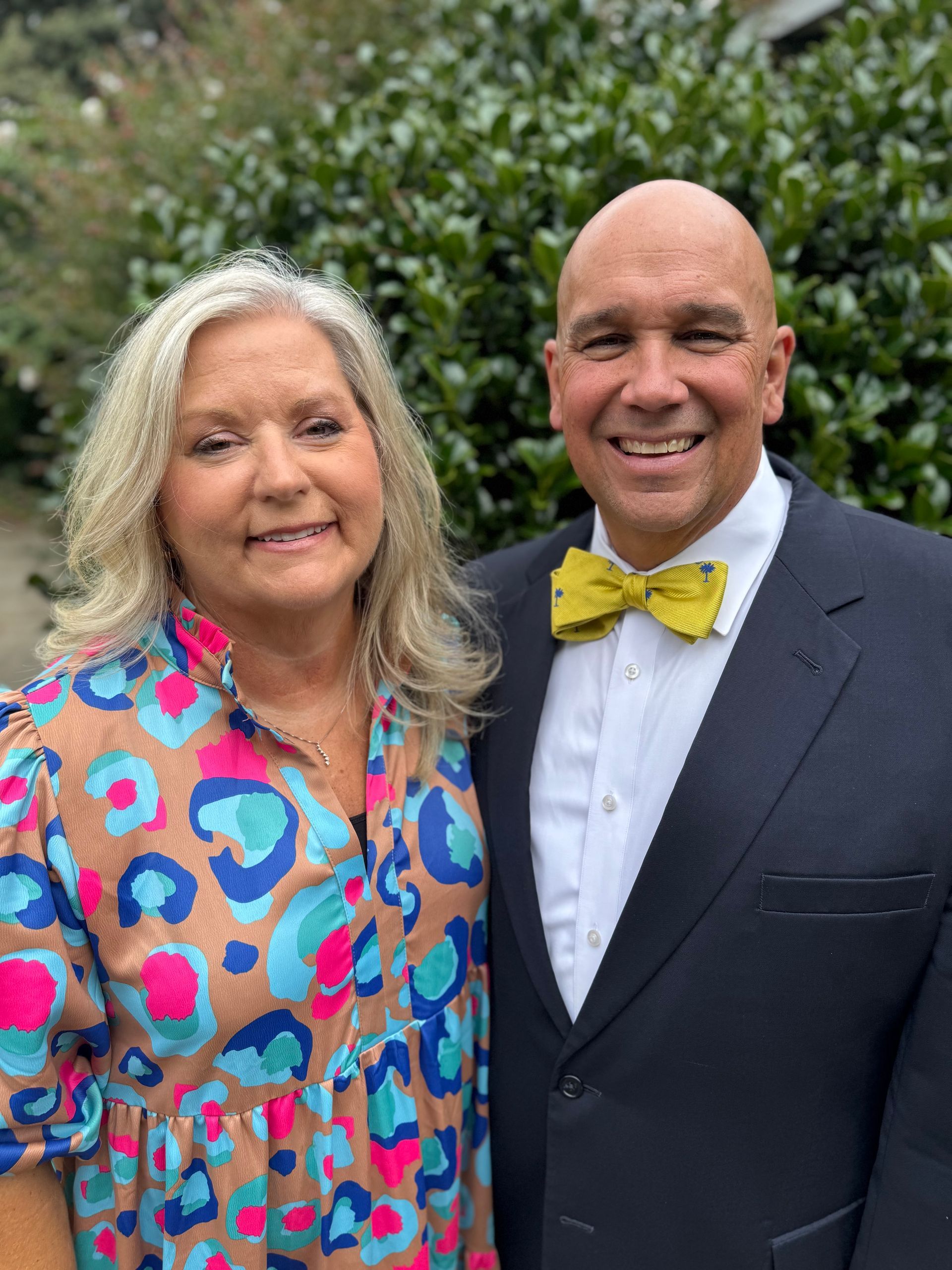 Couple smiling, woman in patterned top, man in suit with yellow bow tie, outdoors.
