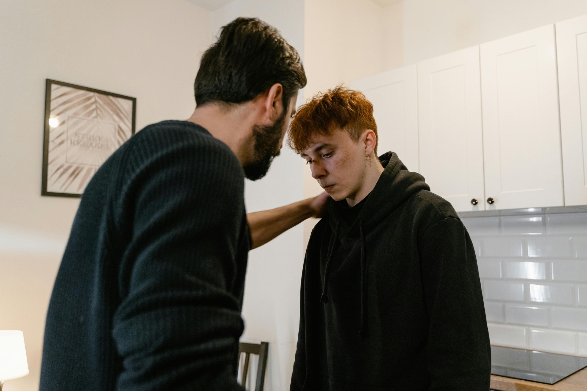 Man consoles a young person indoors. Man has hand on the young person's shoulder. Kitchen setting.