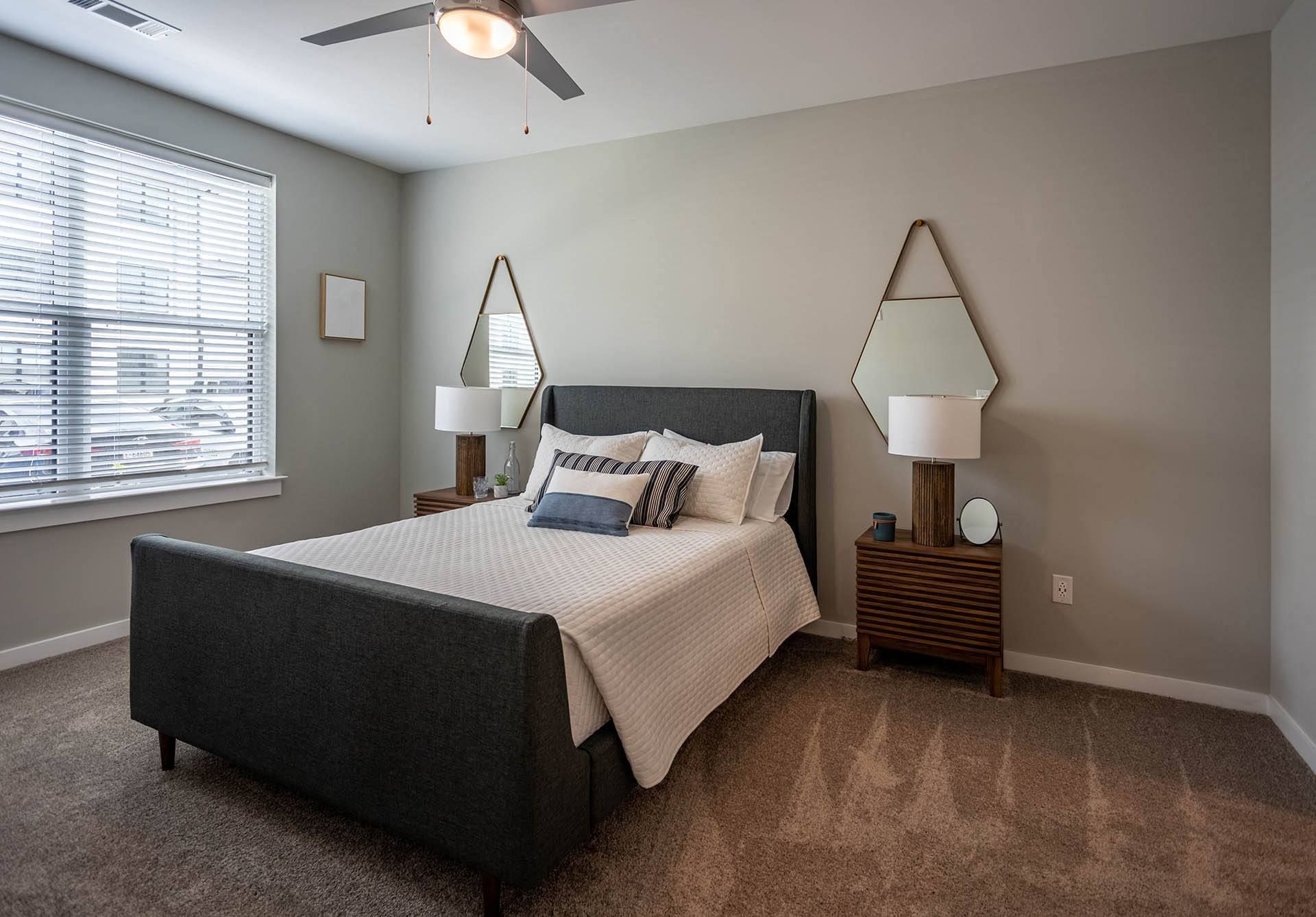 Apartments for Rent in Hixson, TN - Ridgeview at Northgate - Bedroom with Carpeted Floor, Soft Grey Walls, Bed, Nightstands with Lamps, Mounted Mirrors, Ceiling Fan, and Large Window with Blinds.