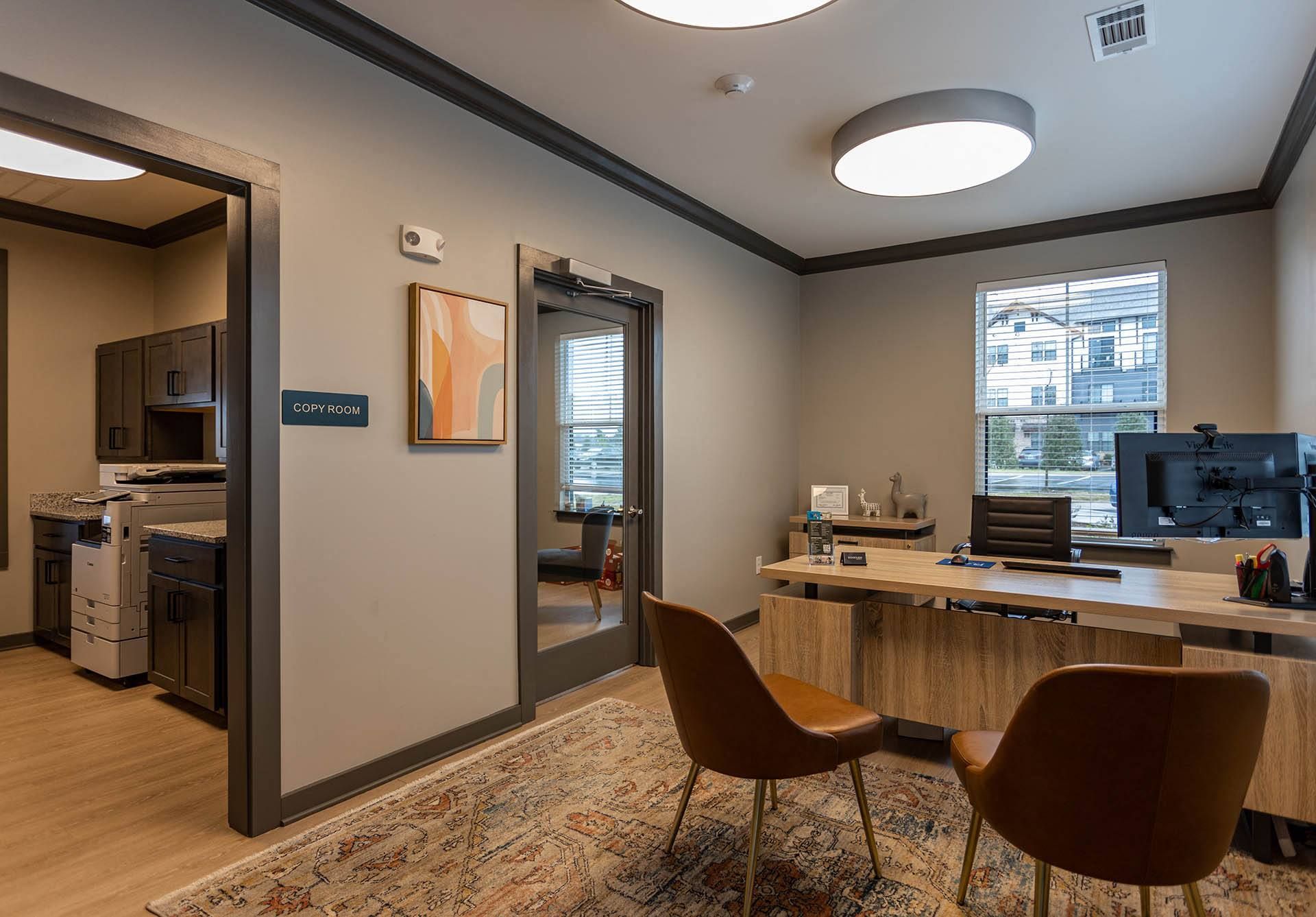 a room with a desk and chairs and a kitchen in the background