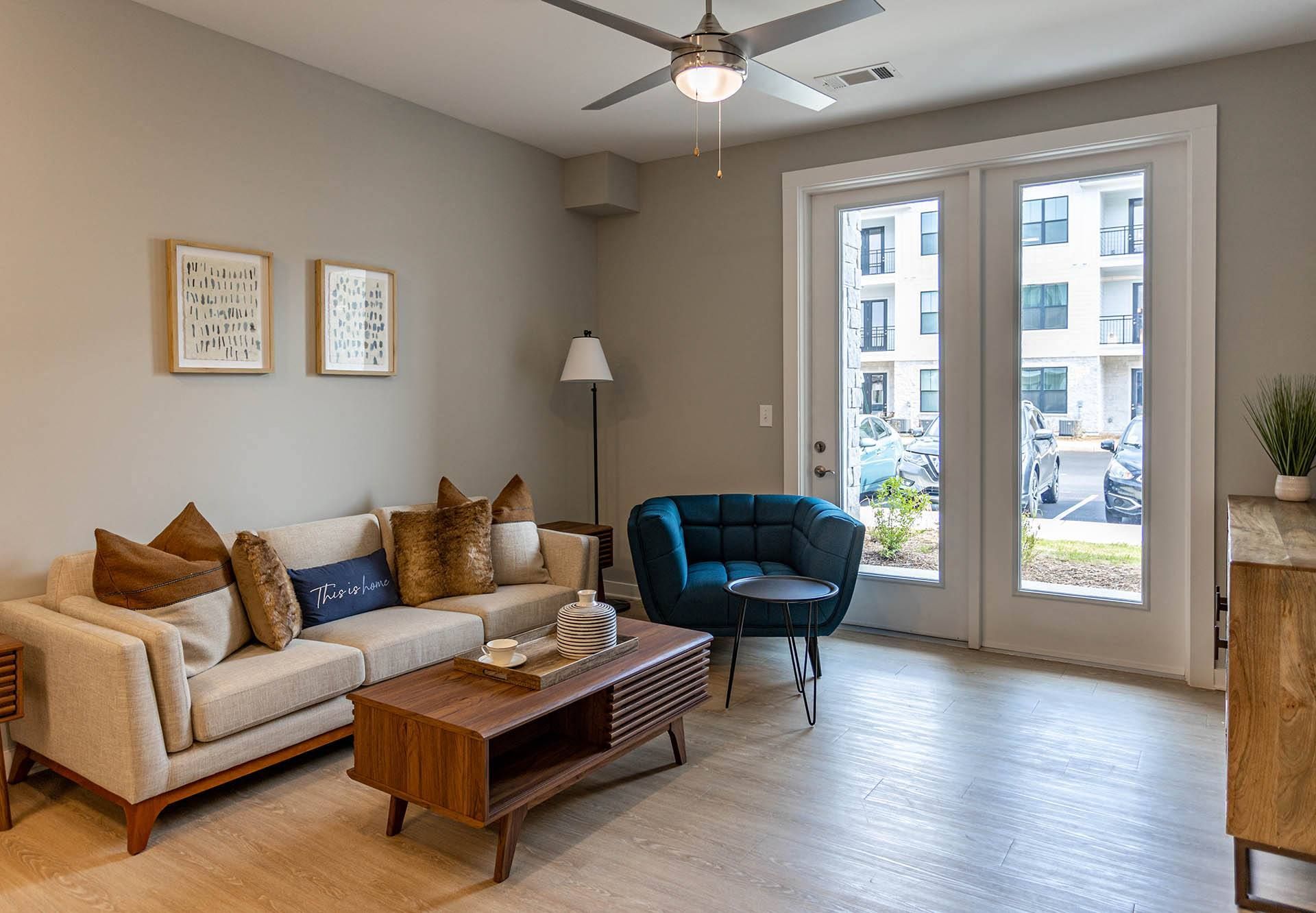 Apartments in Hixson, TN - Ridgeview at Northgate - Living Room with Luxury Vinyl Plank Floor, Beige Sofa, Blue Armchair, Wooden Coffee Table, Side Tables, Floor Lamp, Cabinet, Ceiling Fan, and Glass Doors.
