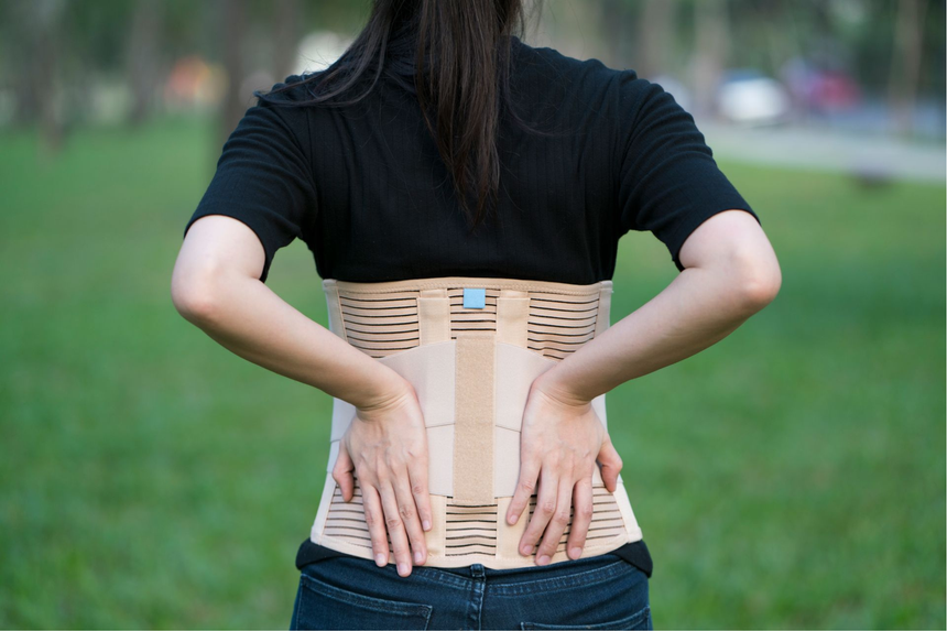 Una persona con indosso un corsetto ortopedico beige si tiene la parte bassa della schiena in un'area erbosa all'aperto.