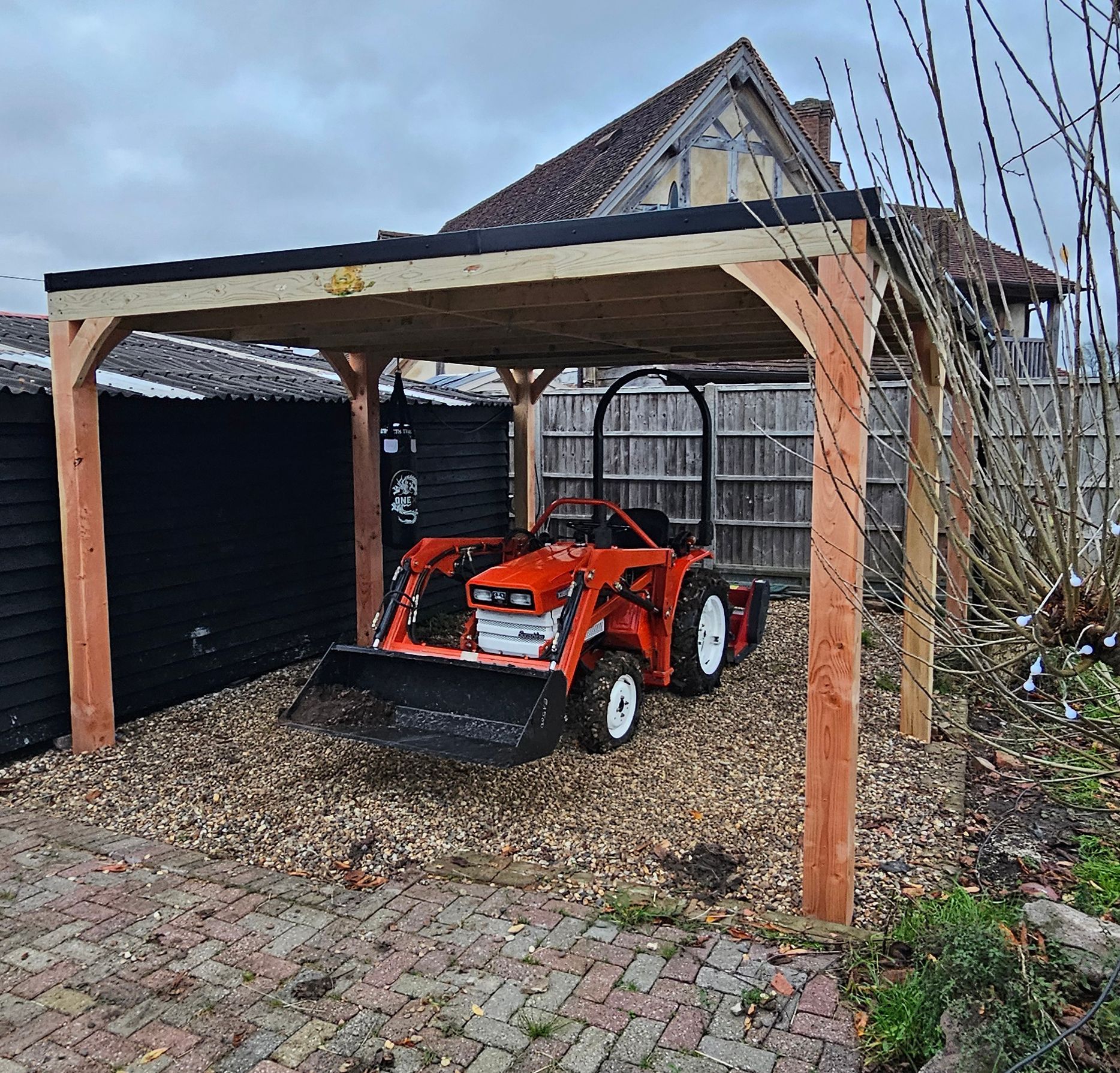 An red compact tractor parked under a wooden carport on a gravel surface next to a dark building.