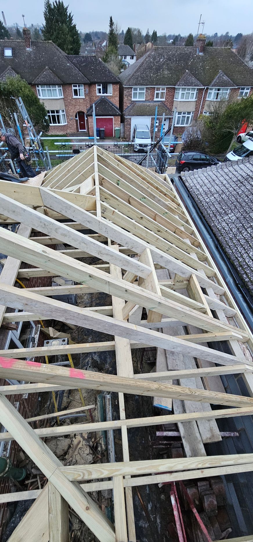 A wooden roof frame under construction on a residential house, viewed from above, with neighbouring homes in the background.