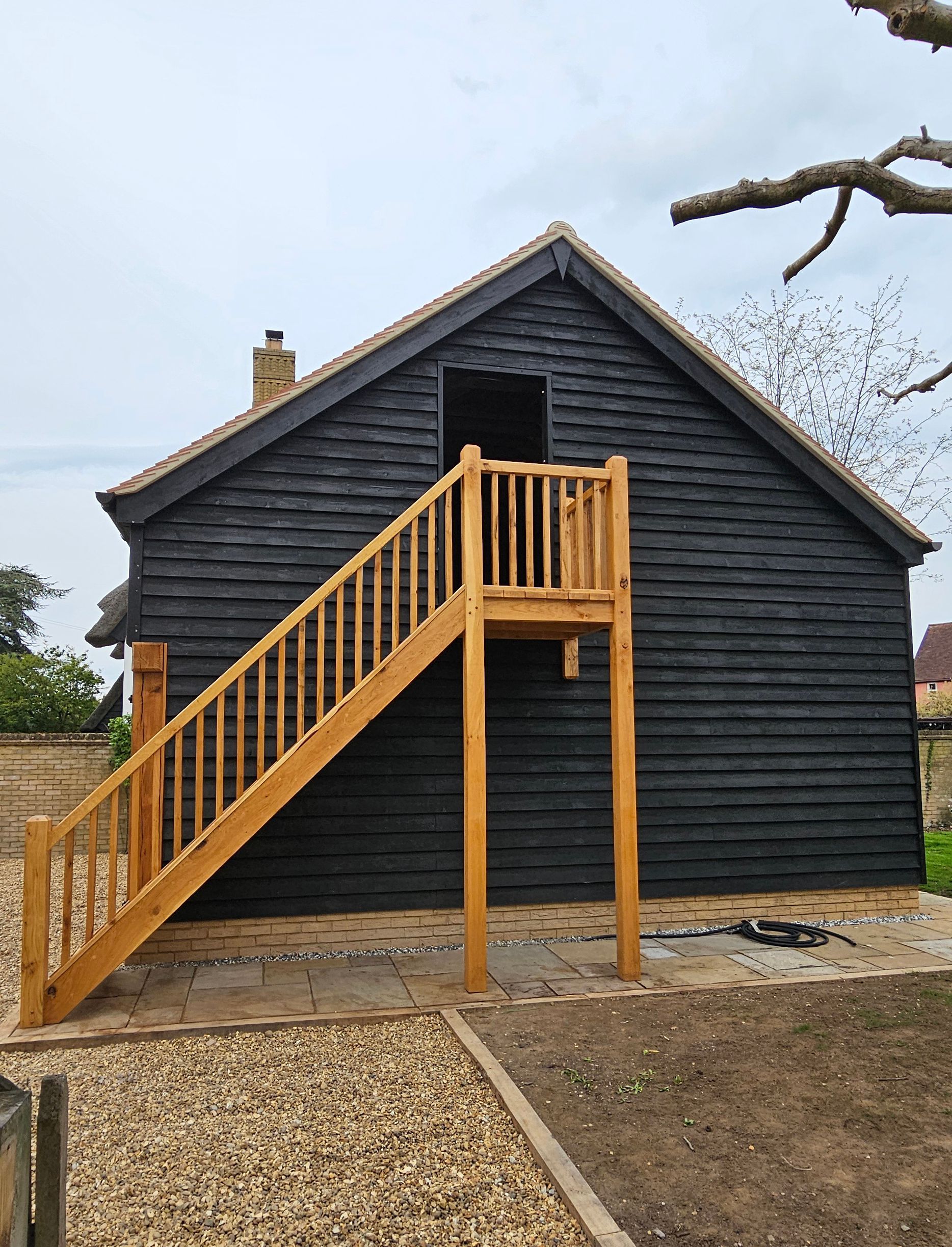 A black, oak framed, timber-clad building features exterior oak stairs leading to an upper-level doorway.