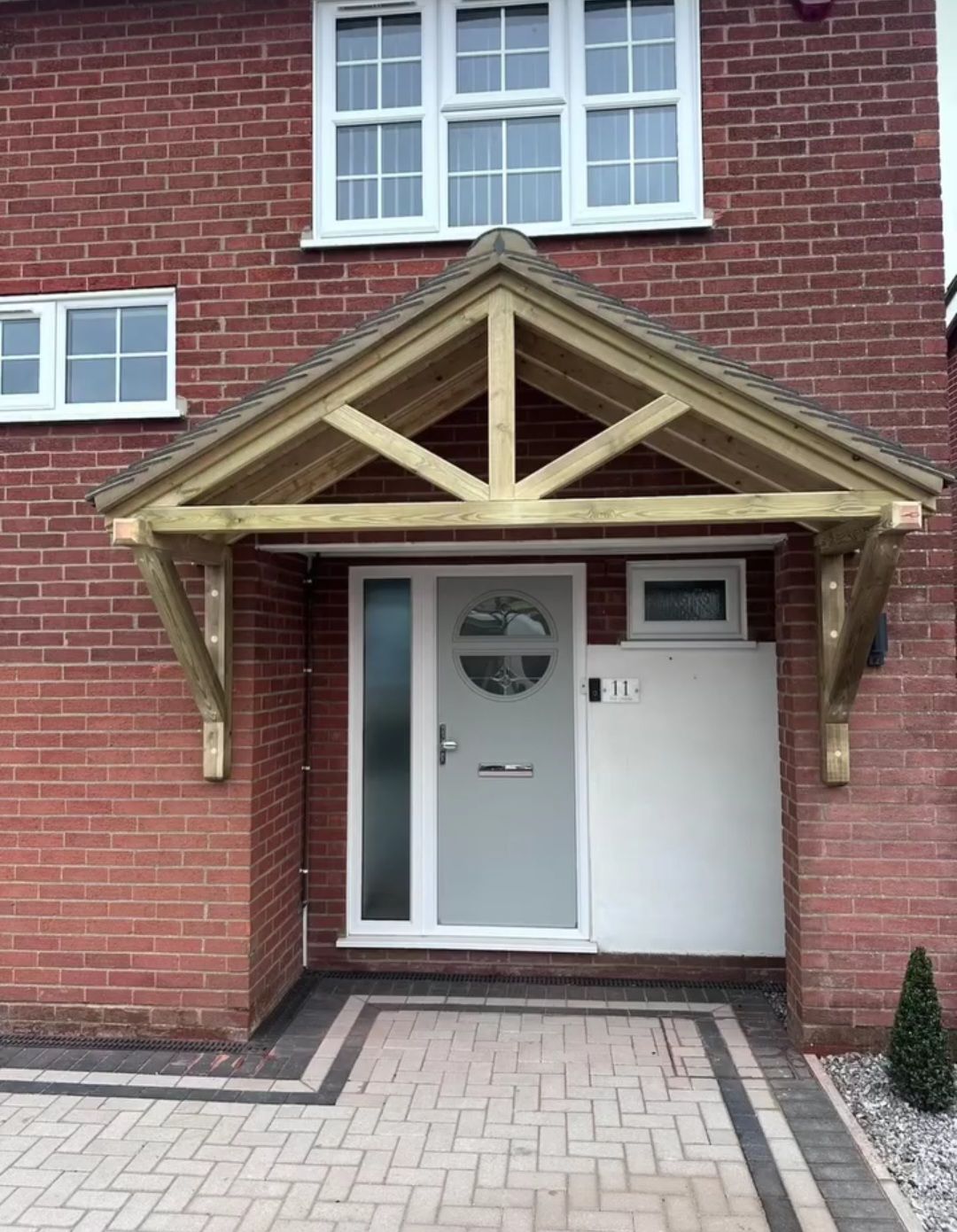 A light grey front door with a circular window under a wooden porch gabled canopy on a red brick house exterior.