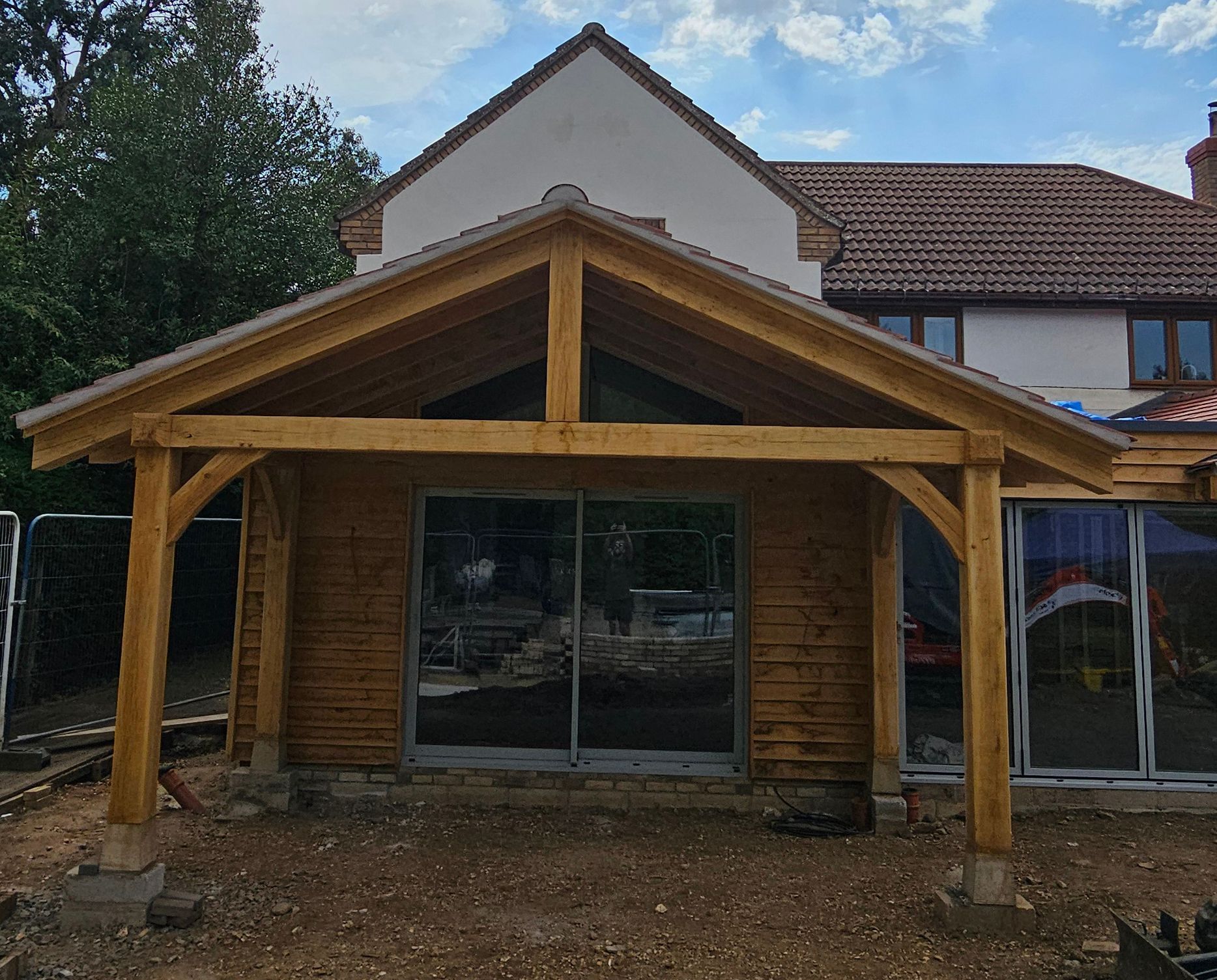 A newly constructed oak timber-frame porch extension attached to the back of a house, set over a gravel construction site.