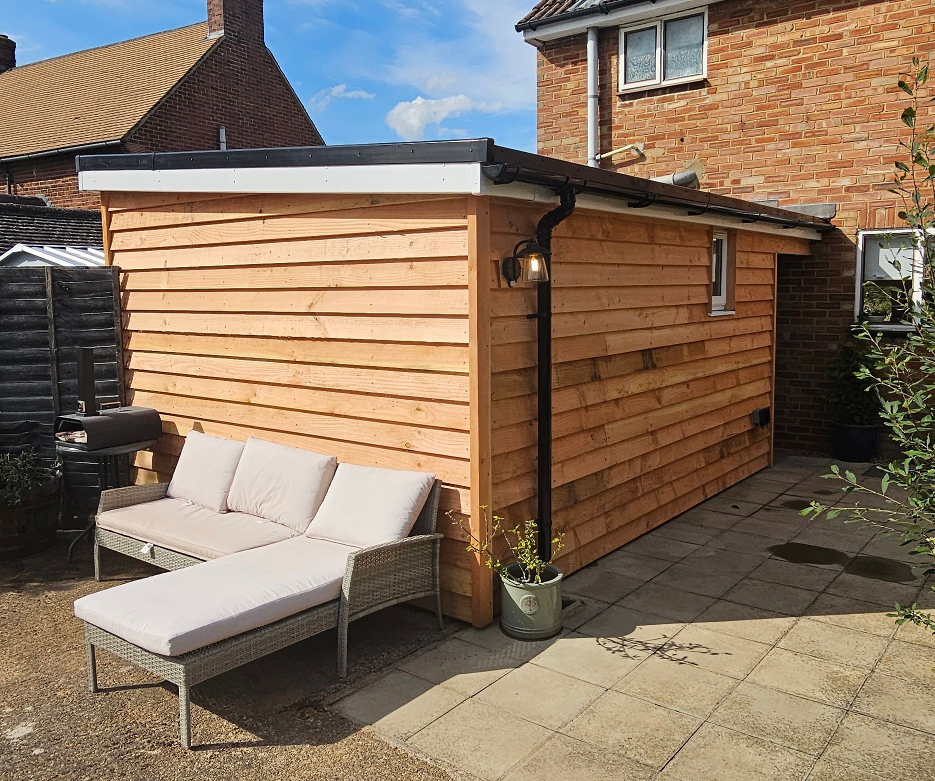 A wooden garden outbuilding with feather edged stands in a backyard next to a brick house.