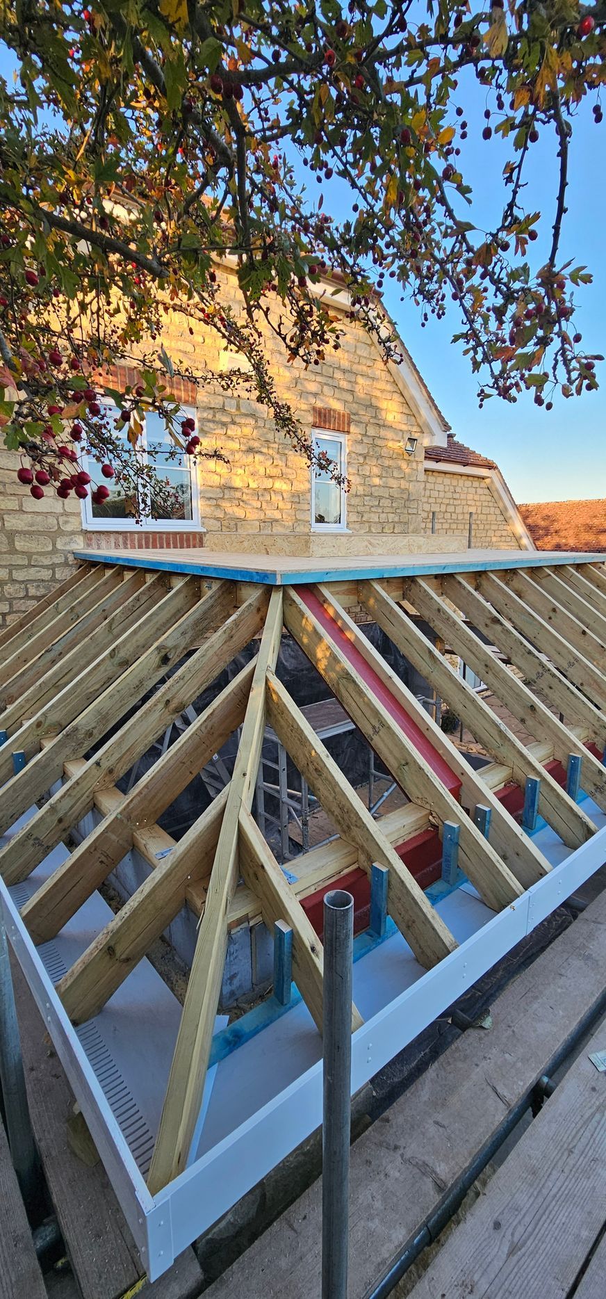 A wooden roof frame for a home extension, viewed from scaffolding against a stone house wall under a blue sky.