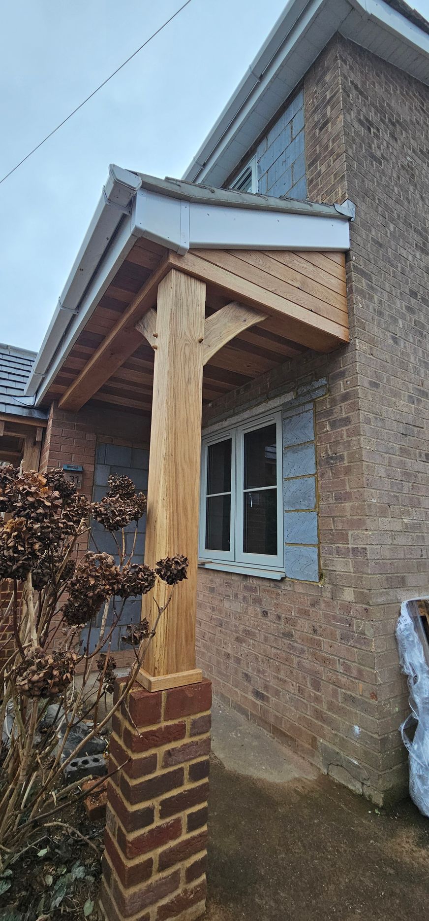 A wooden porch overhang with a single support pillar resting on a brick pier, attached to the side of a brick house.