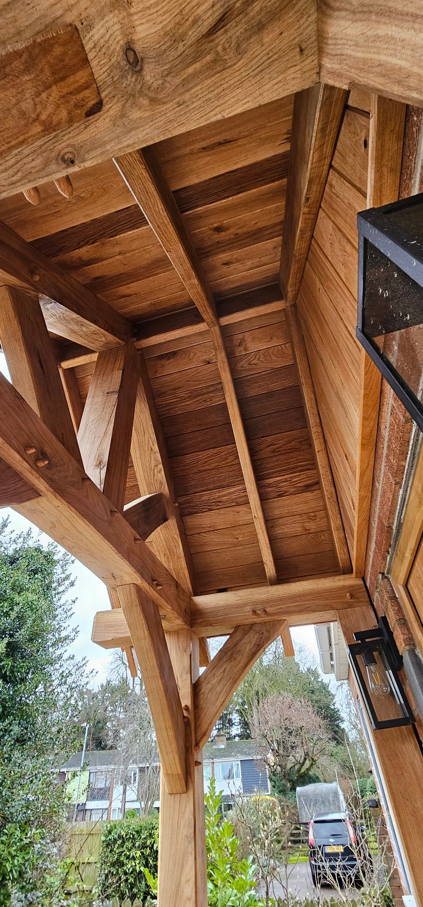 A low-angle view of a rustic oak timber-framed porch ceiling showing exposed wooden beams, rafters, and paneled siding.