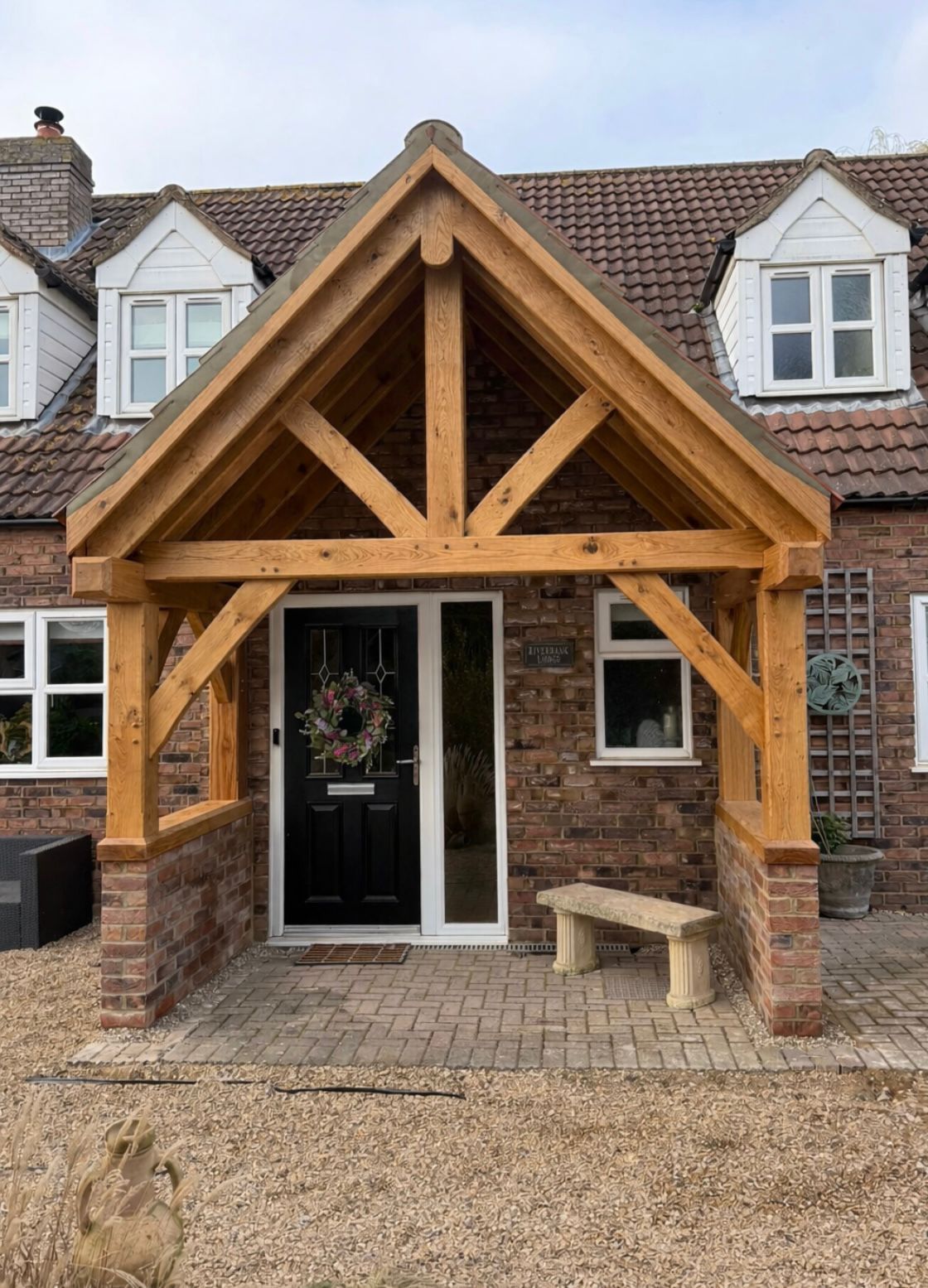 A rustic oak-timbered porch with a pitched roof over a modern black front door, set against a brick house with dormer windows.