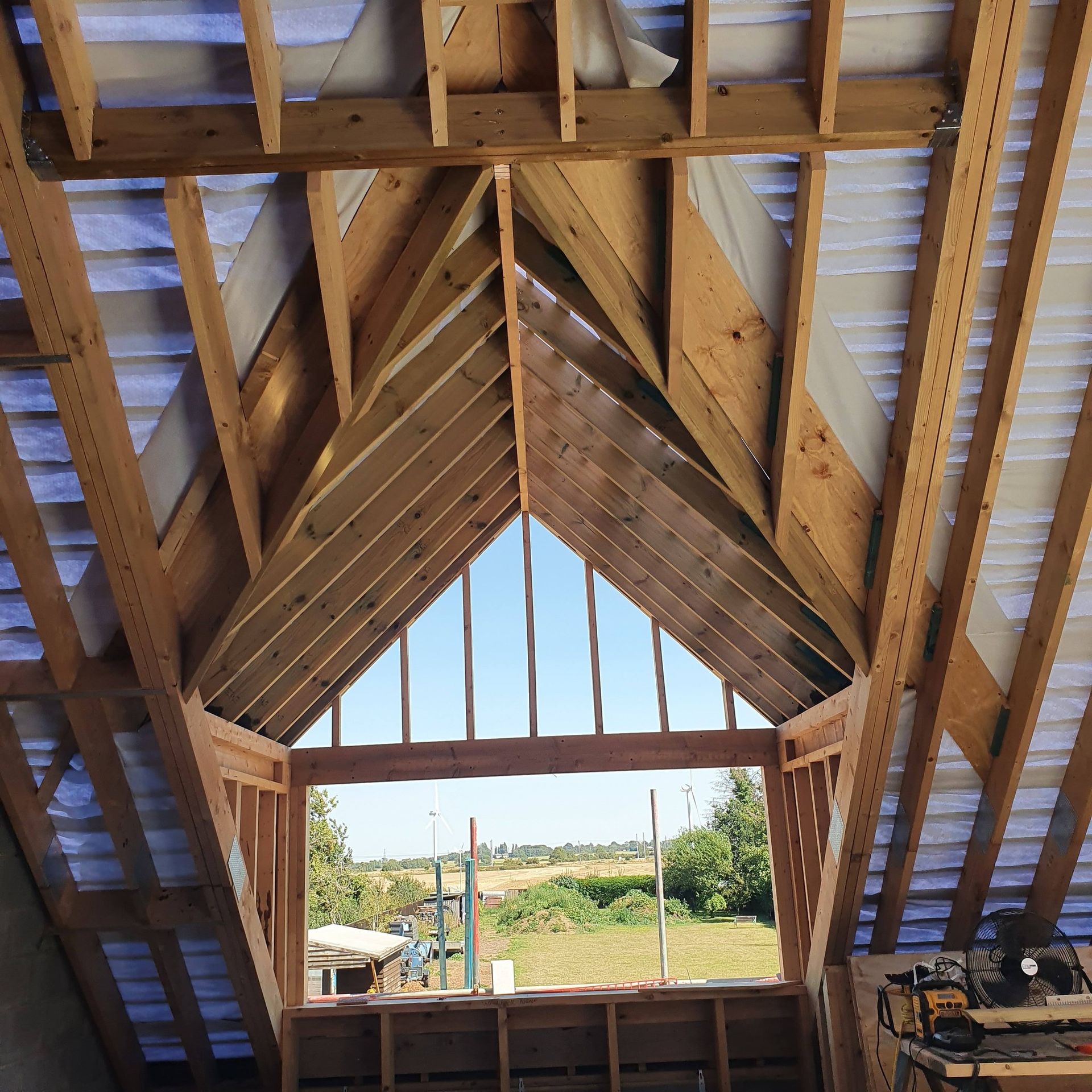 Interior view of a wooden attic frame with a large, triangular gable window opening overlooking a rural landscape.