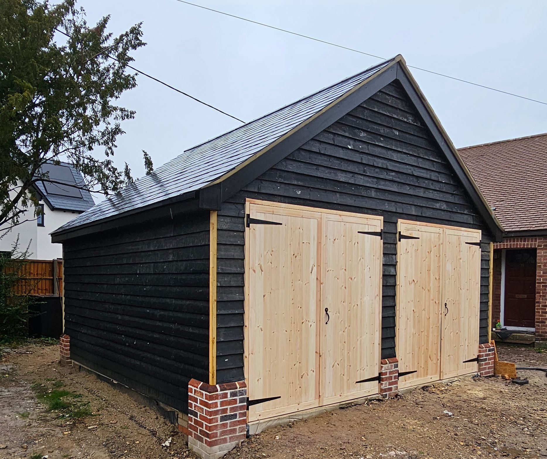 A newly constructed garage with black wooden cladding, two light-wood double doors, and brick corner accents on gravel.