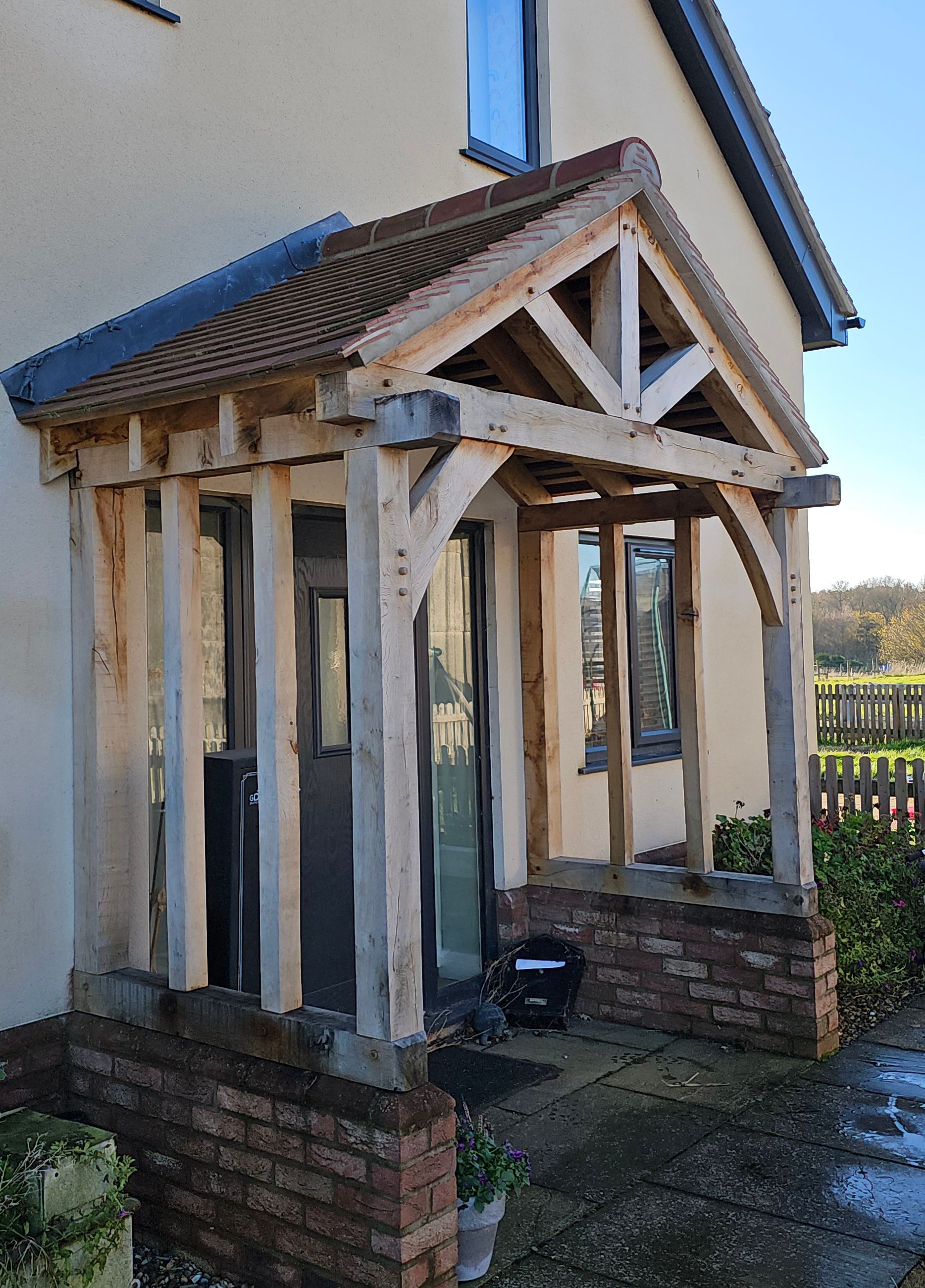 A rustic woak porch frame with a gabled roof, built onto a brick base against the side of a light-colored house.