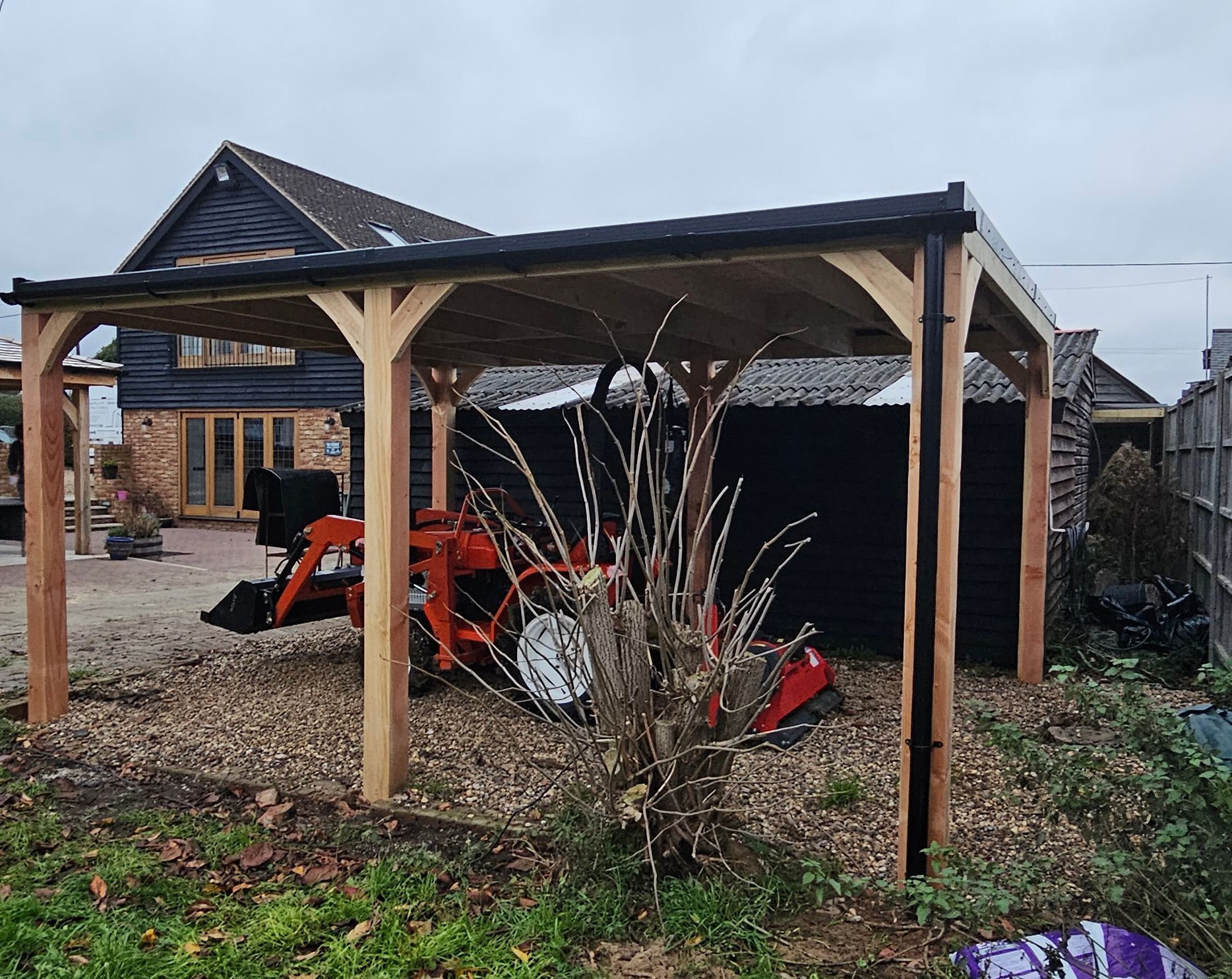 An orange tractor parked under a wooden-framed carport with a black metal roof.