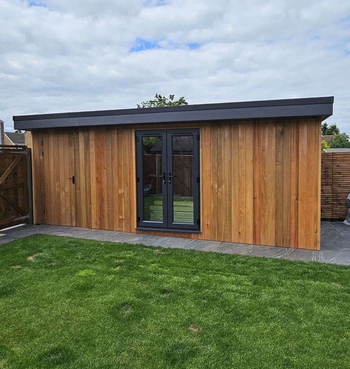 A modern garden room with vertical wood siding and black double doors, set in a backyard with green grass.