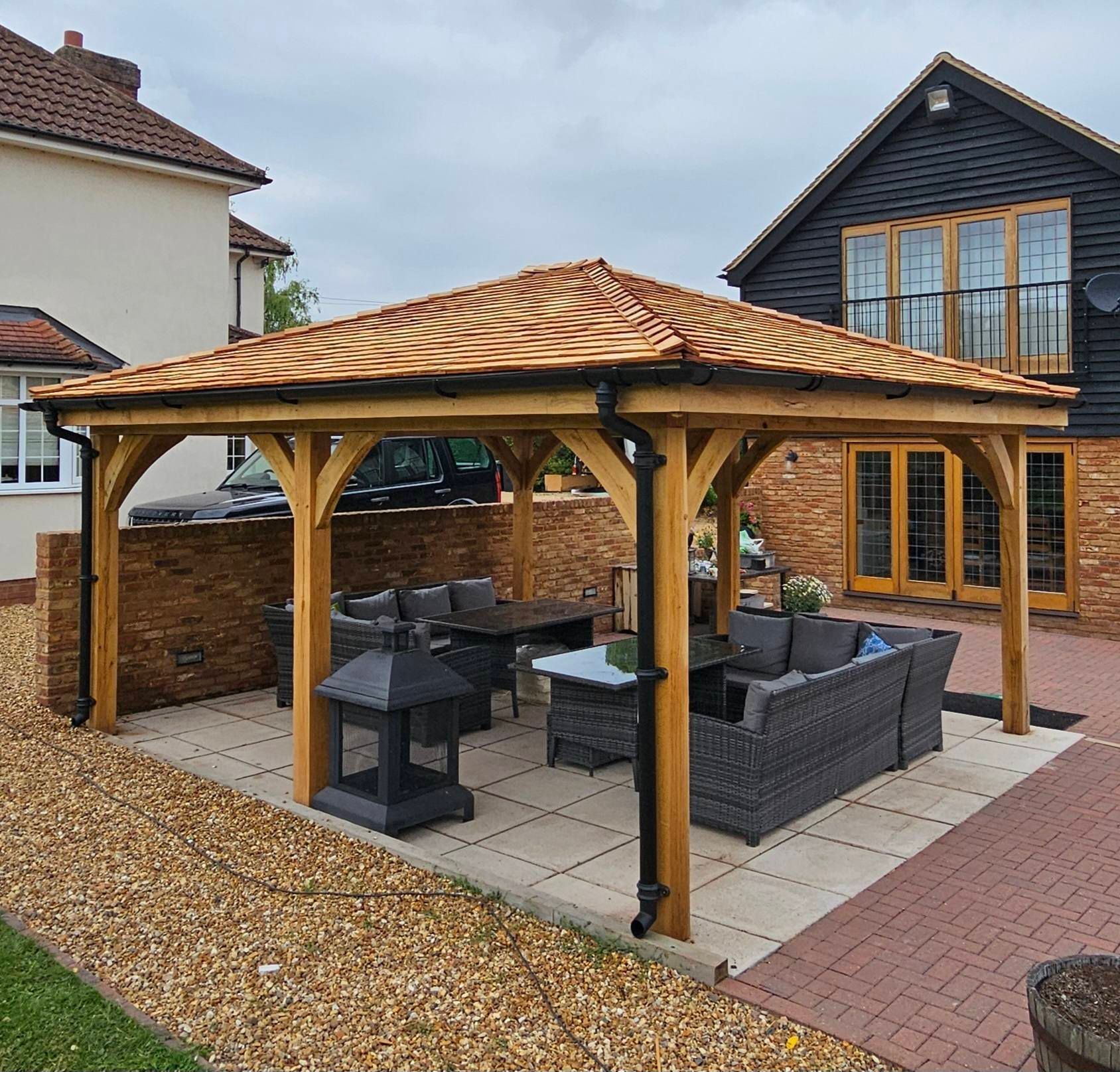Oak outdoor gazebo with a shingle tiled roof sits on a paved patio with grey furniture and a fireplace between two houses.