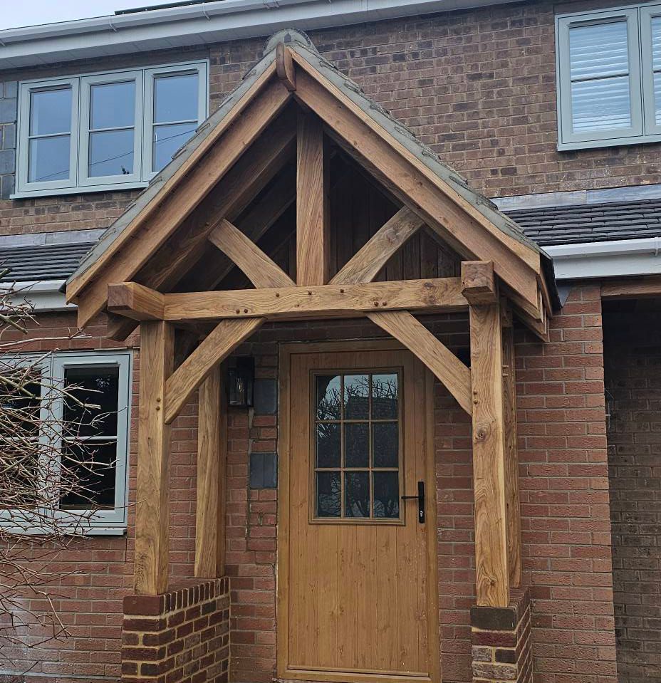 Oak wood-framed front porch canopy with a gabled roof attached to a brick house with gray-framed windows and a wooden door.