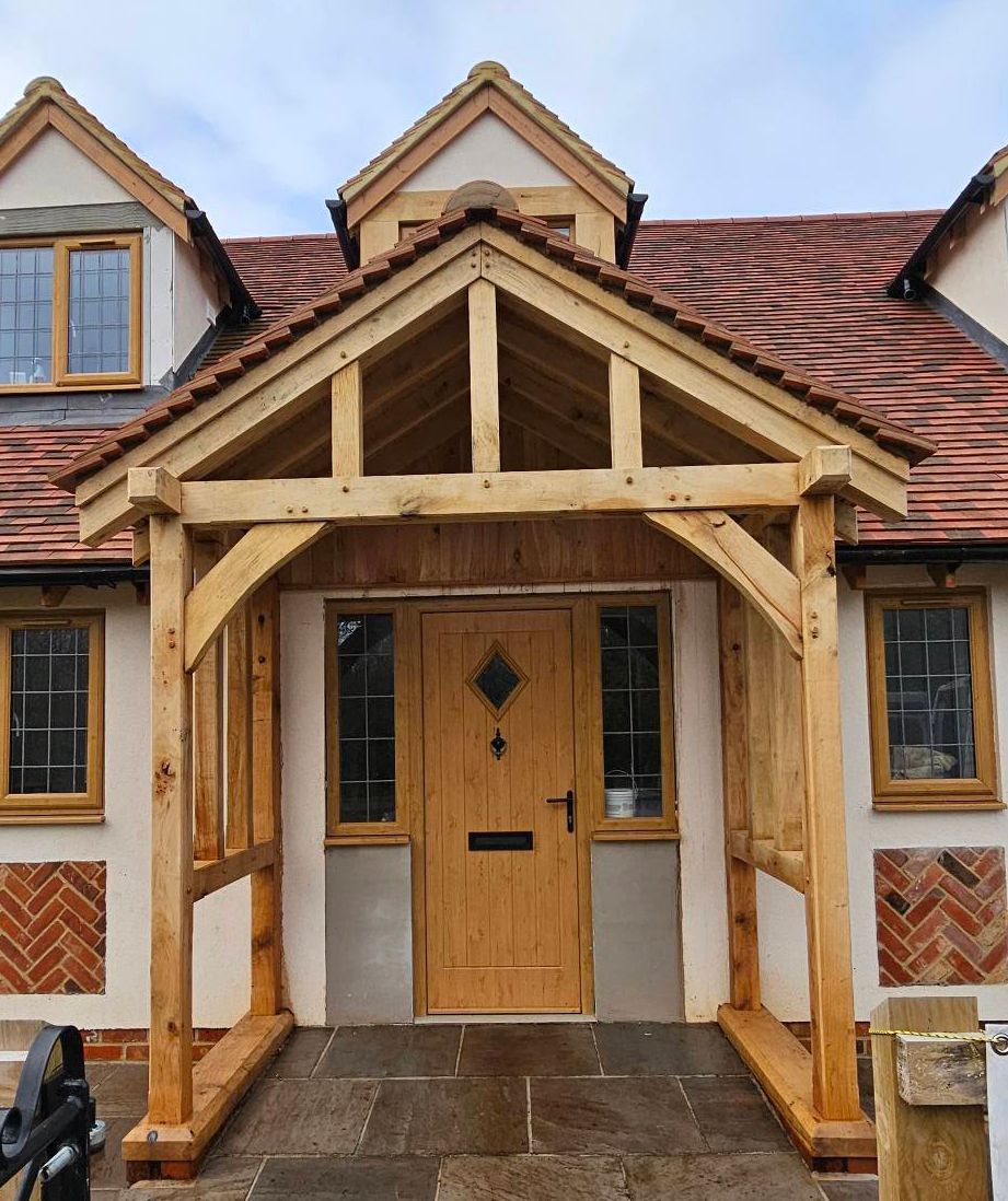 Traditional hand crafted Oak porch entry for a house with a tiled roof, timber frame, and a wooden front door with side windows.