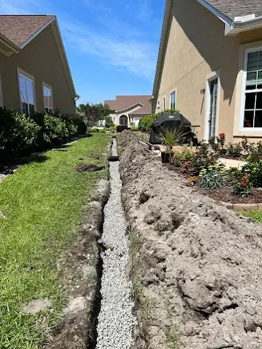 Narrow trench filled with gravel in a backyard between two houses on a sunny day.