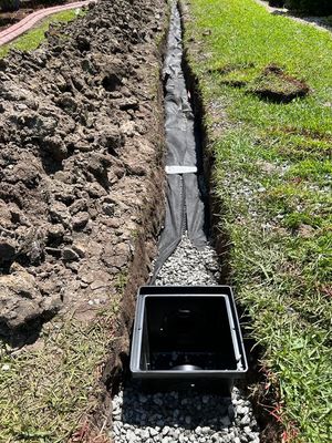 Open trench drain being installed in a yard; black drain, gray gravel, black landscape fabric, dirt, grass.
