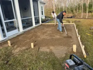 Two people leveling gravel for a patio. A house is on the left, a forest in the background, and a compactor on the right.