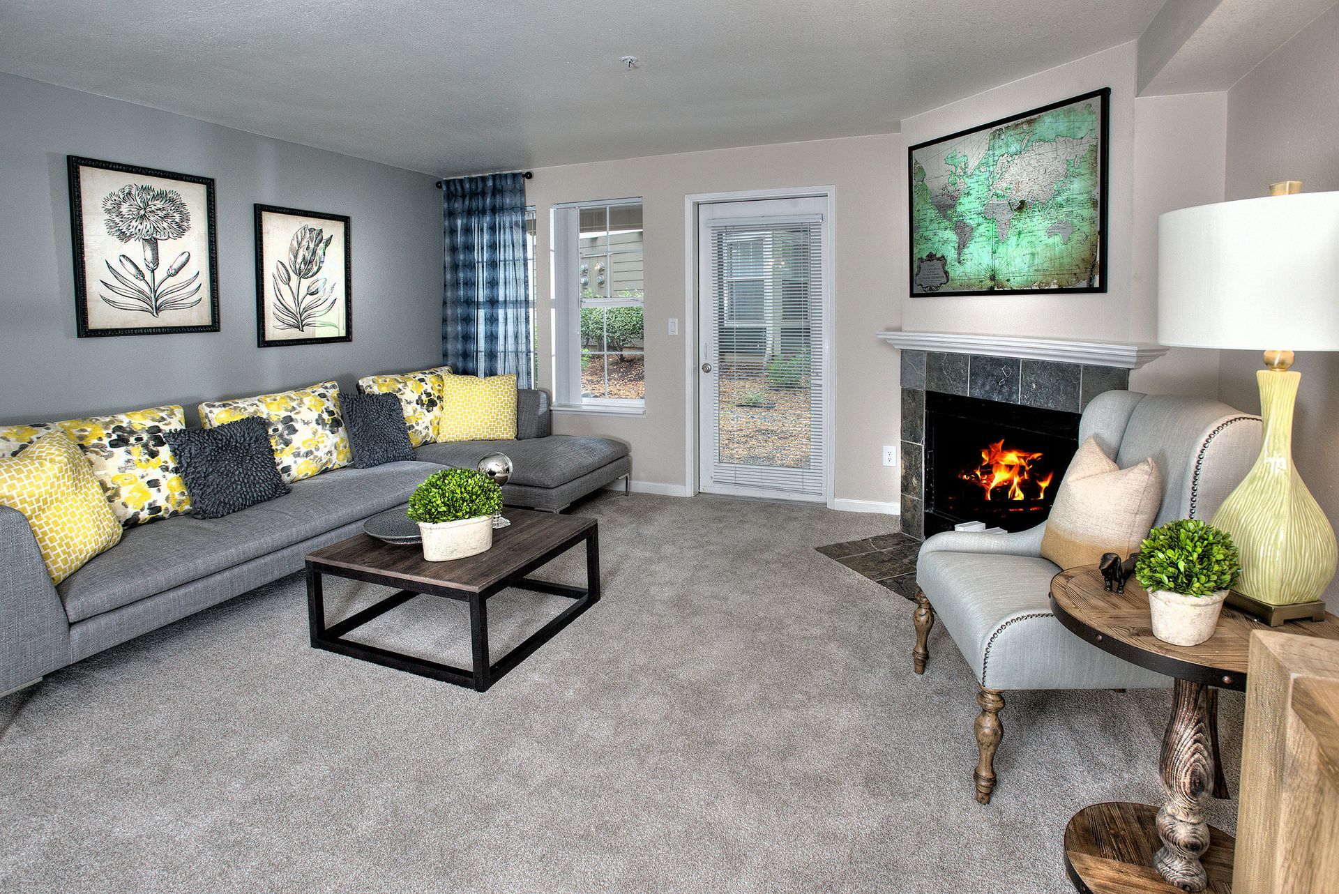 Living room in an apartment with a gray sectional, fireplace, map artwork above the mantel, and a glass door to the outside.