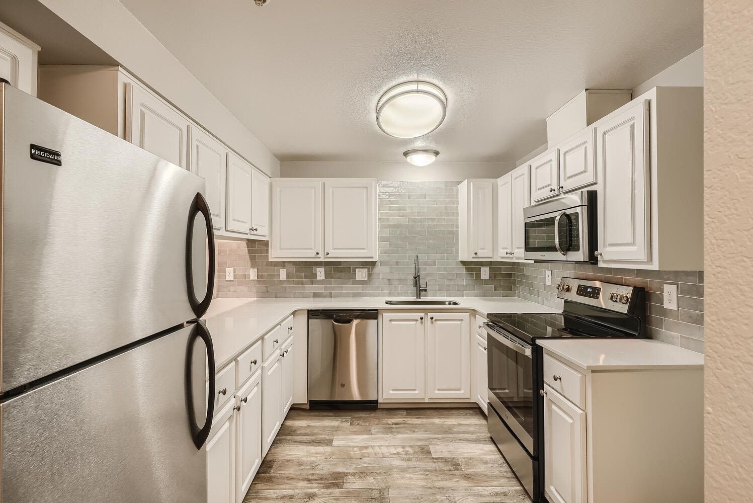 Bright L-shaped kitchen with white cabinets, stainless steel fridge, dishwasher, oven, and tile backsplash.