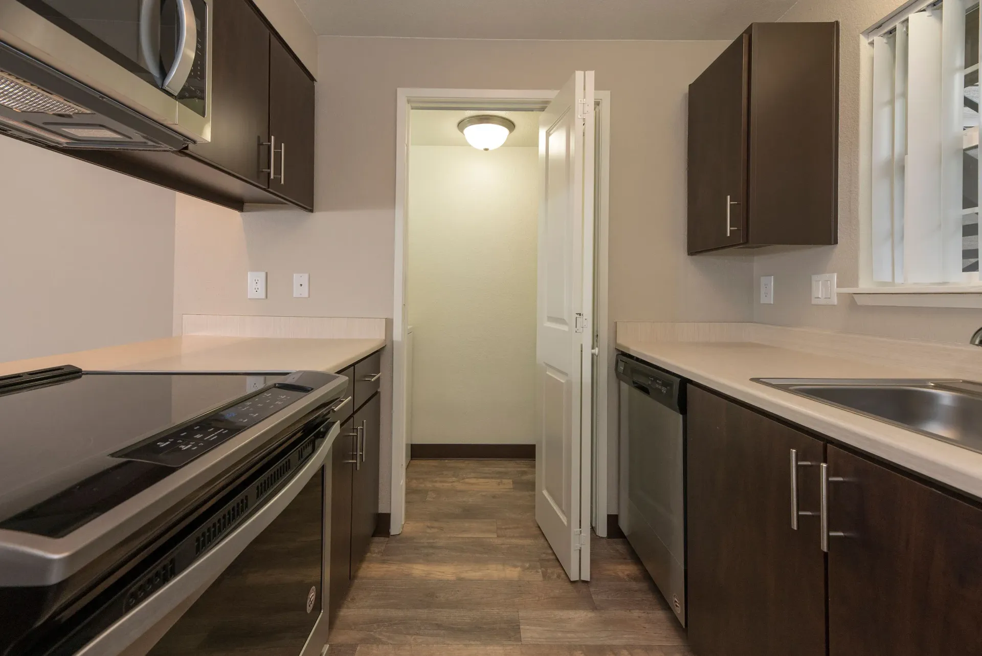 Apartment kitchen with dark cabinets, stainless appliances, and a doorway to a hallway.