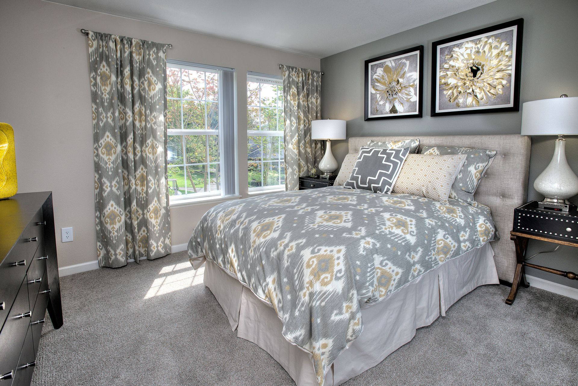 Bedroom featuring a bed with patterned grey bedding, two nightstands, lamps, and a window with patterned curtains.