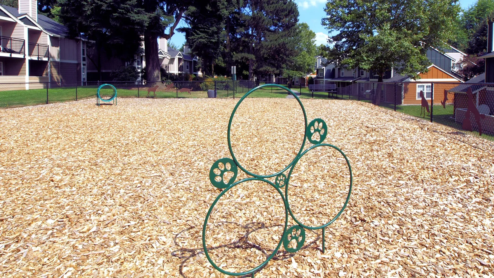 Playground with a green circular climbing frame on wood chips, apartment buildings in the background.