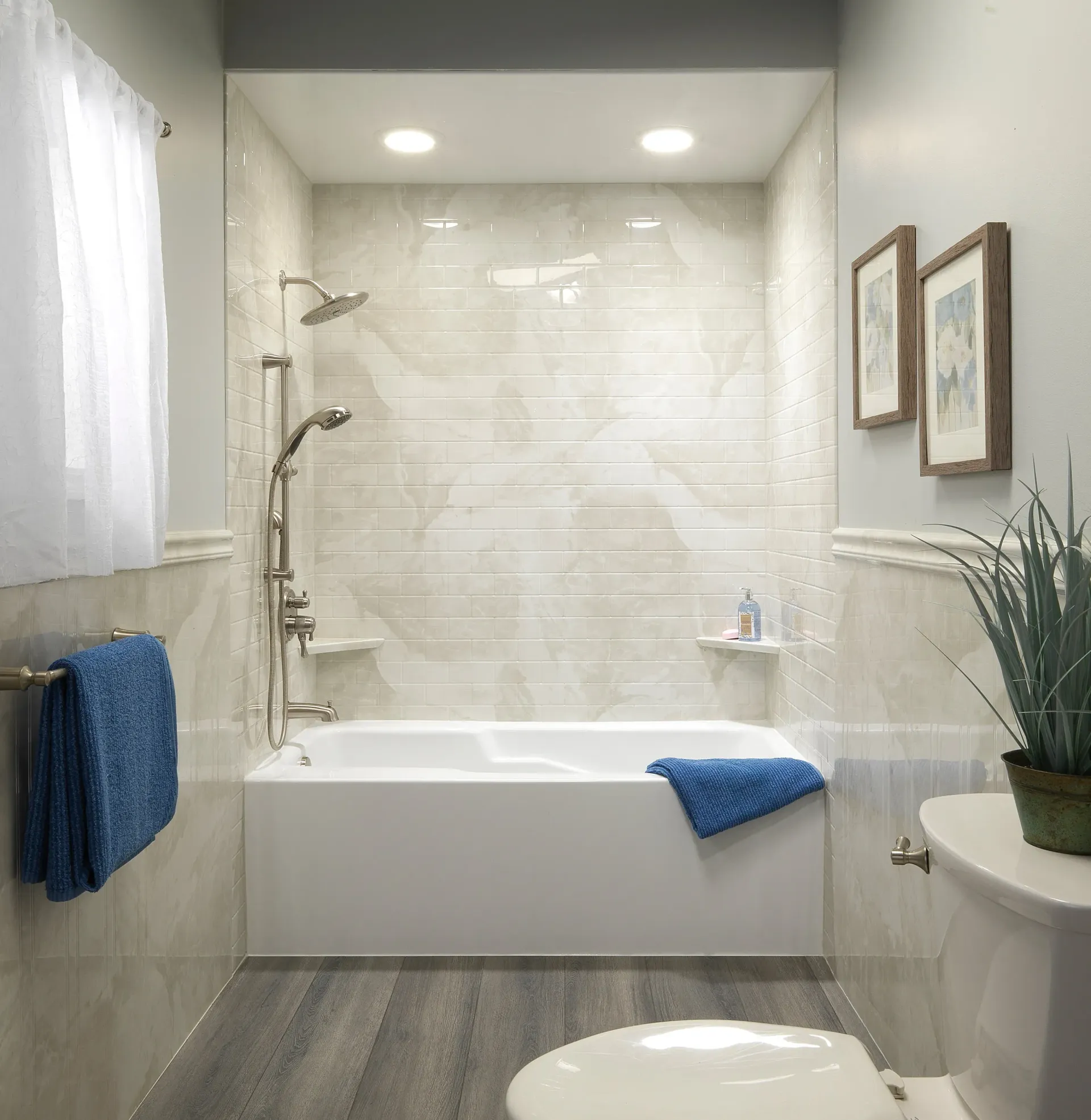 Bright white bathroom with a tub, shower, blue towels, and a toilet beside a plant.