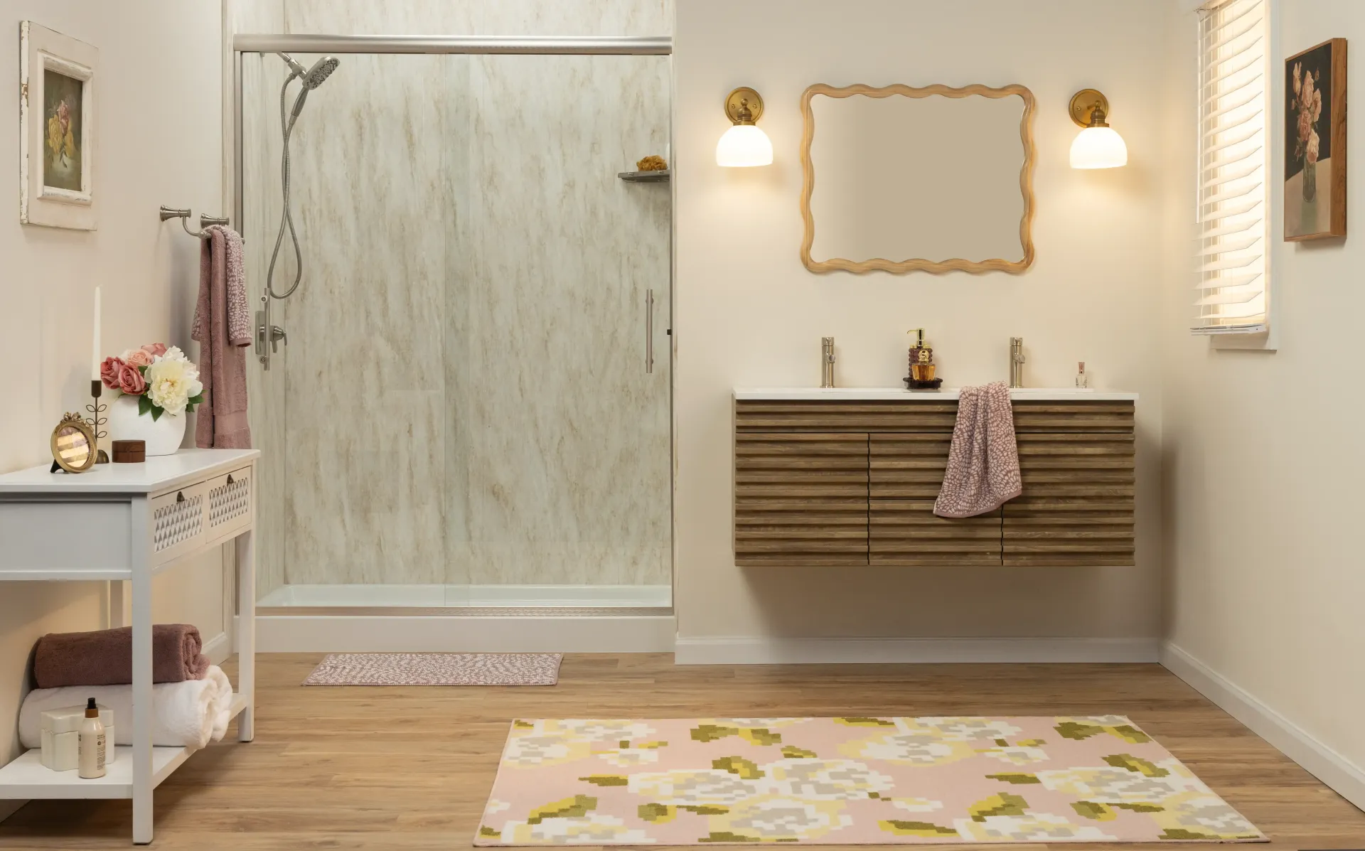 Bright bathroom with glass shower, vanity, gold mirror, wall sconces, and pink floral rug.