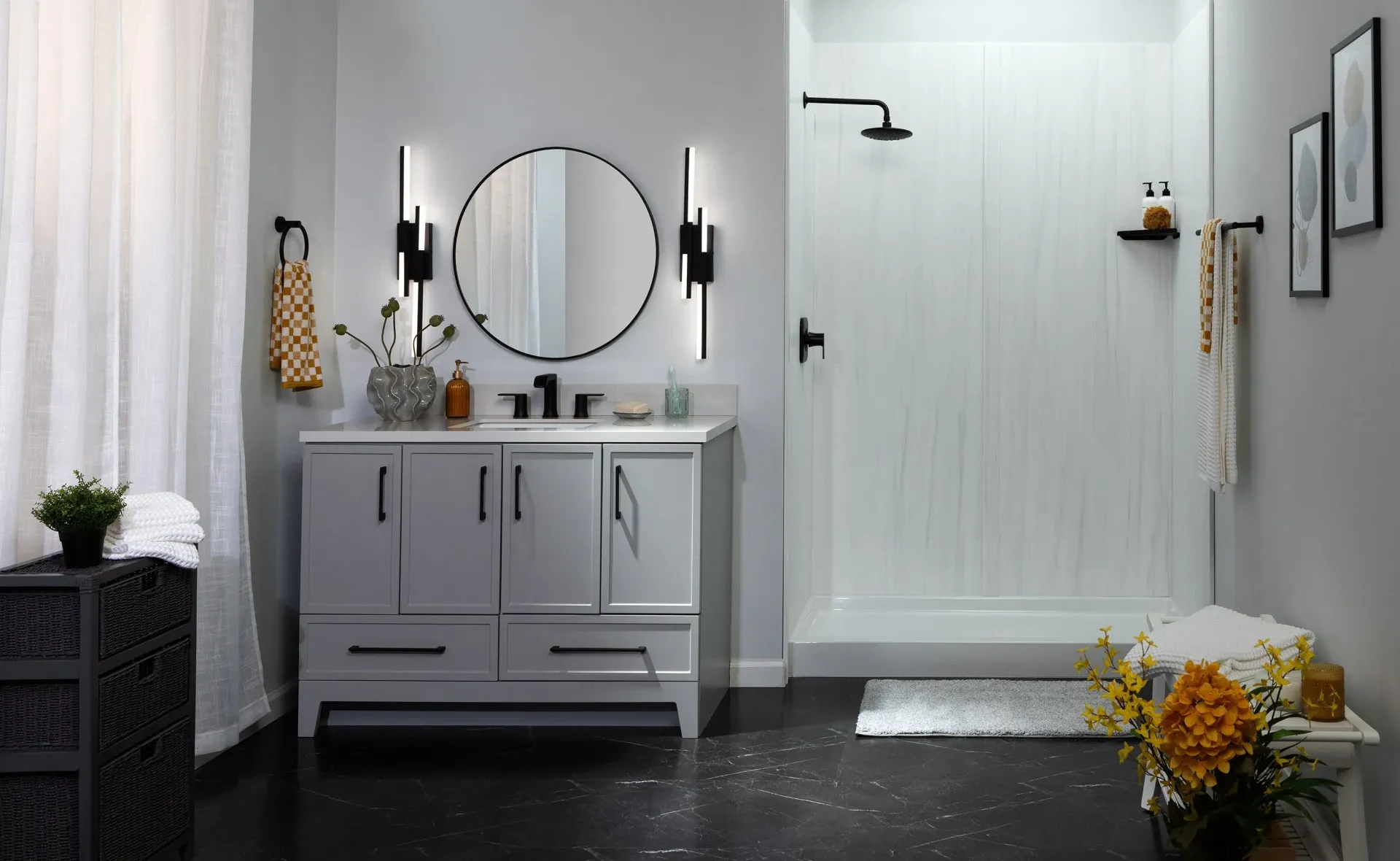 Modern gray bathroom with vanity, round mirror, glass shower, black floor, and small plants.