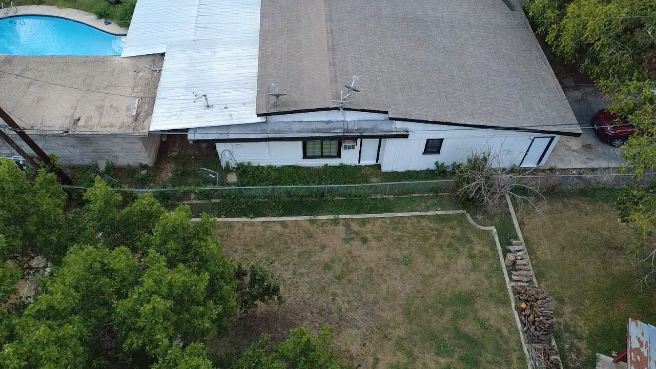 Aerial view of a white building with brown and silver roofs, a swimming pool, and a grassy yard.