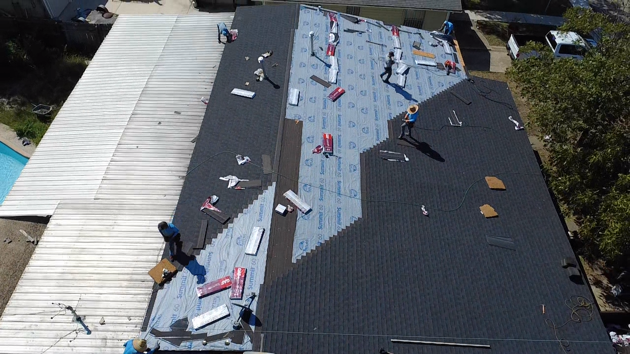 Roofers working on a residential roof, installing dark shingles. Construction tools, materials, & safety equipment visible.