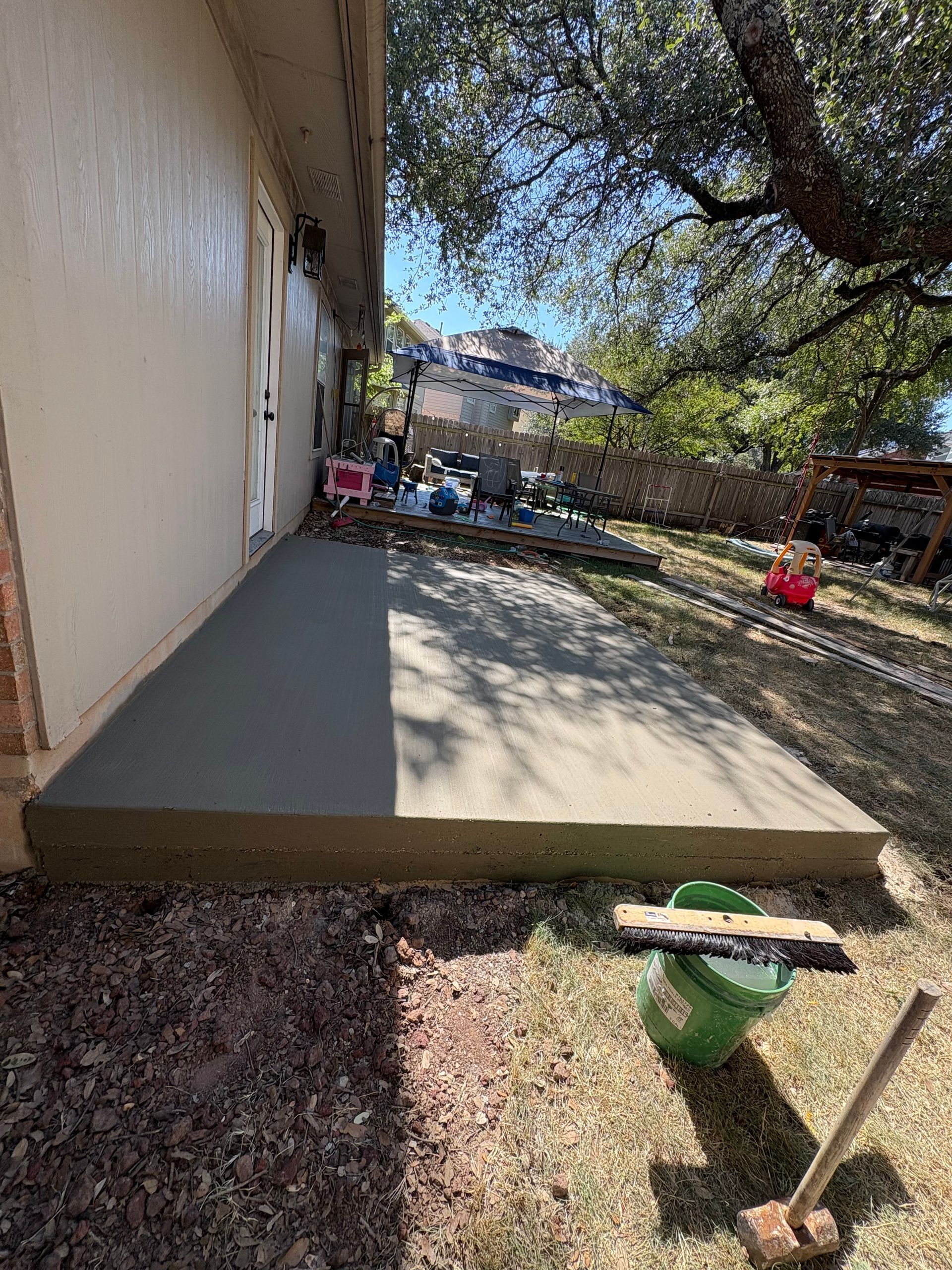 Newly poured concrete patio next to a house with a green bucket and tools in the foreground, sunny day.